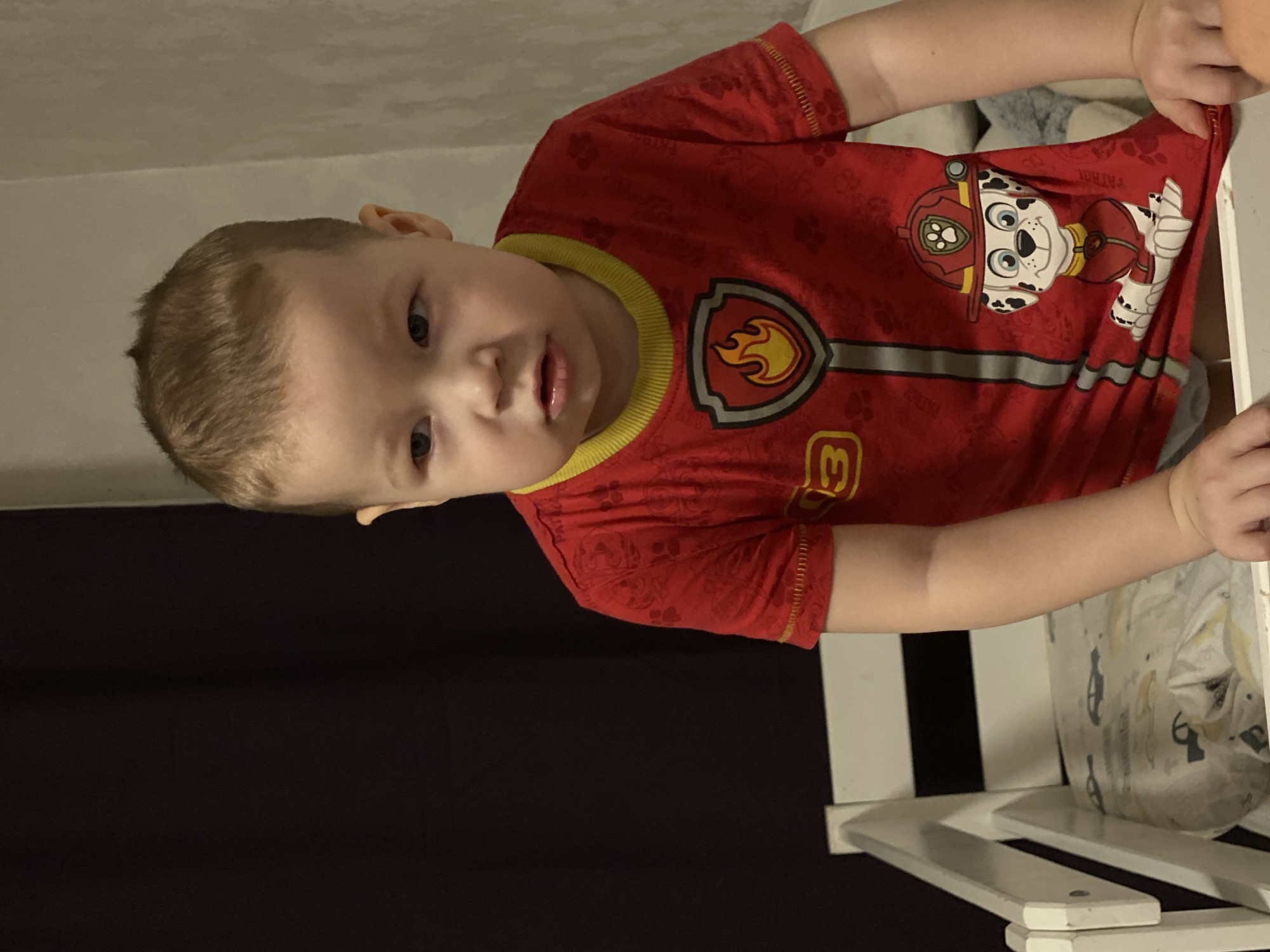 An image taken indoors in Sochi, Russia, depicts a young child, appearing to be a toddler or preschooler, positioned in the foreground. The child has light brown, short-cropped hair and light-colored eyes, directed slightly to the viewer's left with a neutral expression and slightly parted lips. The child is wearing a red short-sleeved t-shirt featuring a yellow ribbed collar. The shirt's design mimics a firefighter's uniform, with a gray shield containing a yellow and orange flame emblem on the upper chest, a vertical gray stripe down the center, a yellow number "3" on the left side, and a large cartoon image of Marshall the Dalmatian firefighter from Paw Patrol on the lower right. The child's hands rest on the top rail of a white wooden bed frame, visible in the lower foreground, which contains light-colored bedding with faint patterns. The background on the left side of the frame is a dark, solid color, likely a curtain or wall, while the background on the right is a lighter, textured surface, possibly a wall or draped curtain. The scene is illuminated by ambient indoor lighting.