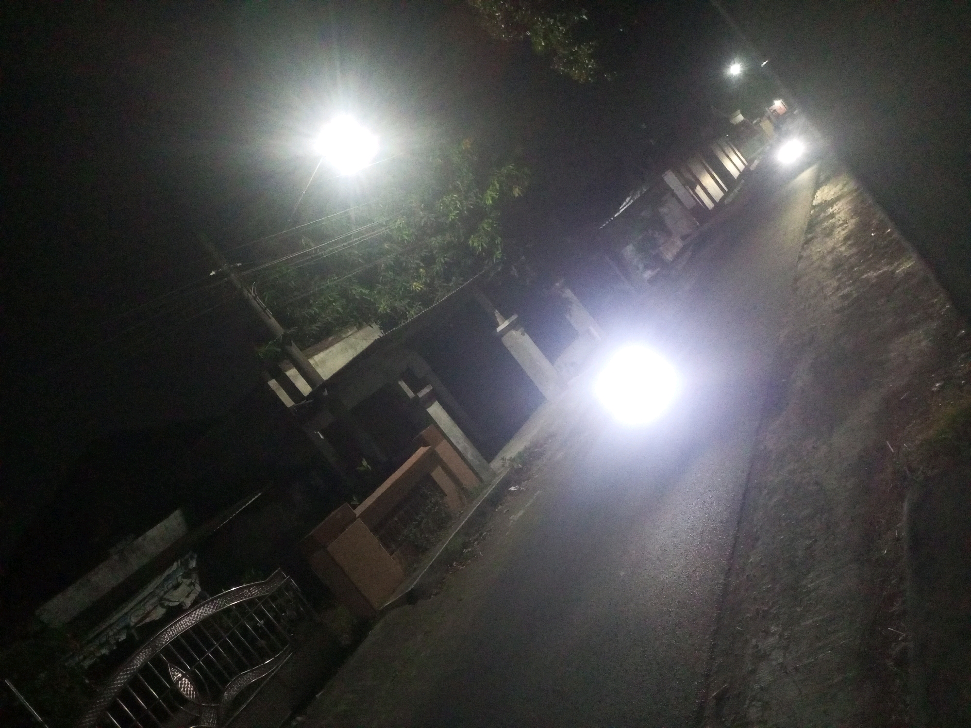 The image depicts a street scene at night, likely in Ngasem, Indonesia, illuminated by streetlights and vehicle headlights. The perspective is from a slightly elevated, angled viewpoint, looking down a paved road bordered by buildings and vegetation.
In the foreground, a metal fence or railing with a decorative pattern is visible on the left. The road surface appears wet and reflective, suggesting recent rain. On the right side of the road, a textured surface, possibly a sidewalk or unpaved area, is visible.
Further down the road, a vehicle's bright headlights illuminate the asphalt. The surrounding environment includes buildings with visible architectural elements like pillars and windows. Trees and foliage are present, partially obscuring some structures. Power lines are also visible in the upper left portion of the image. The overall ambiance is dark, with strong light sources creating bright flares and deep shadows.