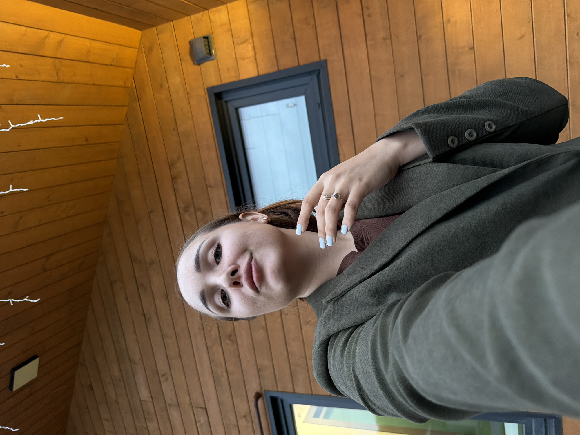 A young woman with light skin and dark hair pulled back is taking a selfie outdoors in front of a wooden building. She is wearing a dark green blazer over a brown shirt. Her left hand is resting on her chest, showing light blue nail polish and a ring on her left index finger. The building has horizontal wooden siding and a window with a dark frame. There are string lights hanging from the eaves of the building. The overall lighting suggests it is late afternoon or early evening. There is no indication of the specific location within Kamyennoye, Russia.