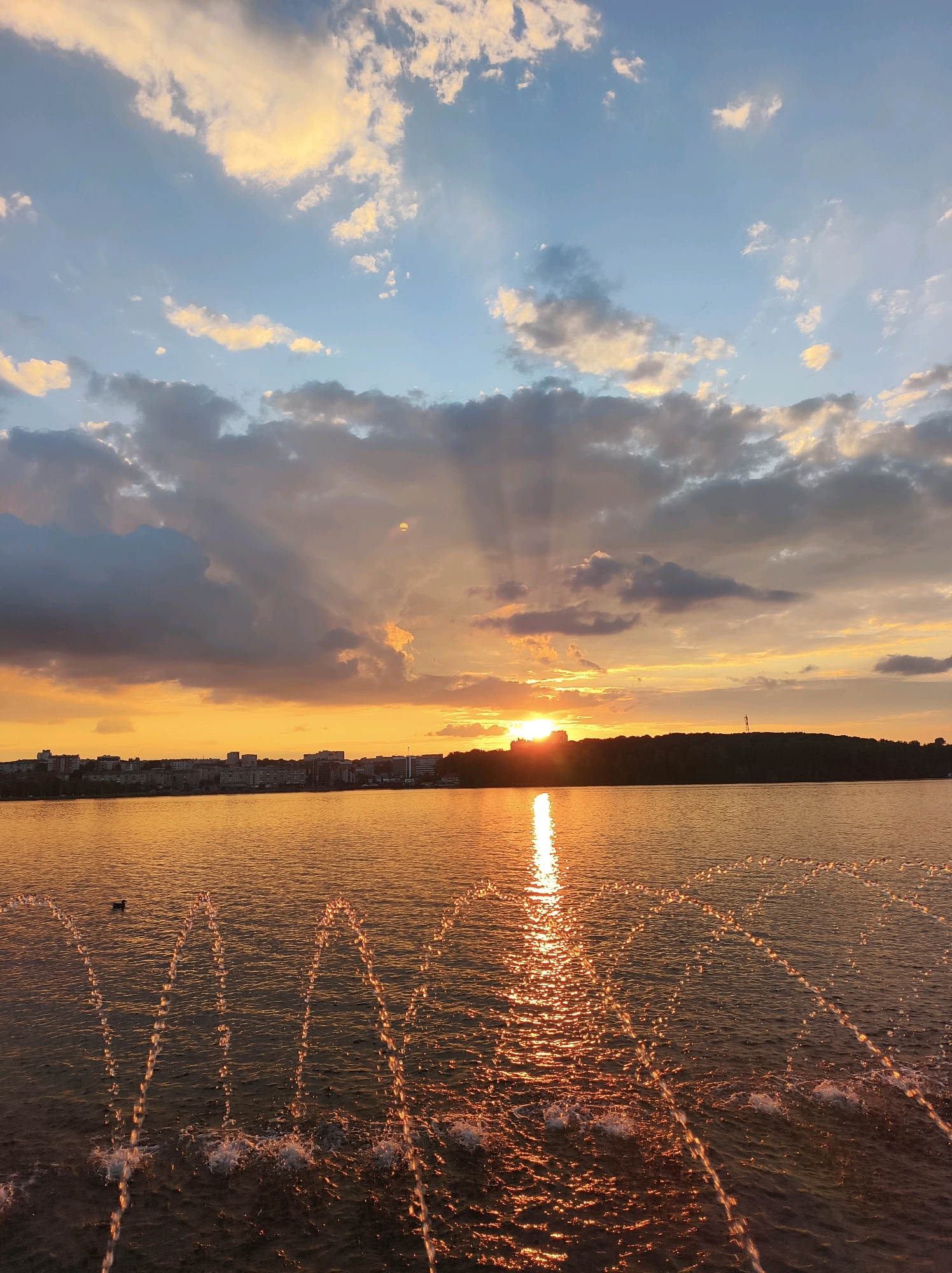 This is a photograph of a sunset over Lake Ternopil in Ternopil, Ukraine. The sky is filled with dramatic clouds, illuminated by the setting sun which casts a golden glow over the scene. Rays of sunlight pierce through the clouds, creating a dramatic effect. In the foreground, water jets from fountains create arc-shaped patterns on the surface of the lake, reflecting the warm colors of the sunset. Across the lake, a treeline and the silhouette of a city are visible on the horizon. A single duck is swimming on the left side of the frame. The overall mood is serene and picturesque, capturing the beauty of a sunset over an urban body of water.