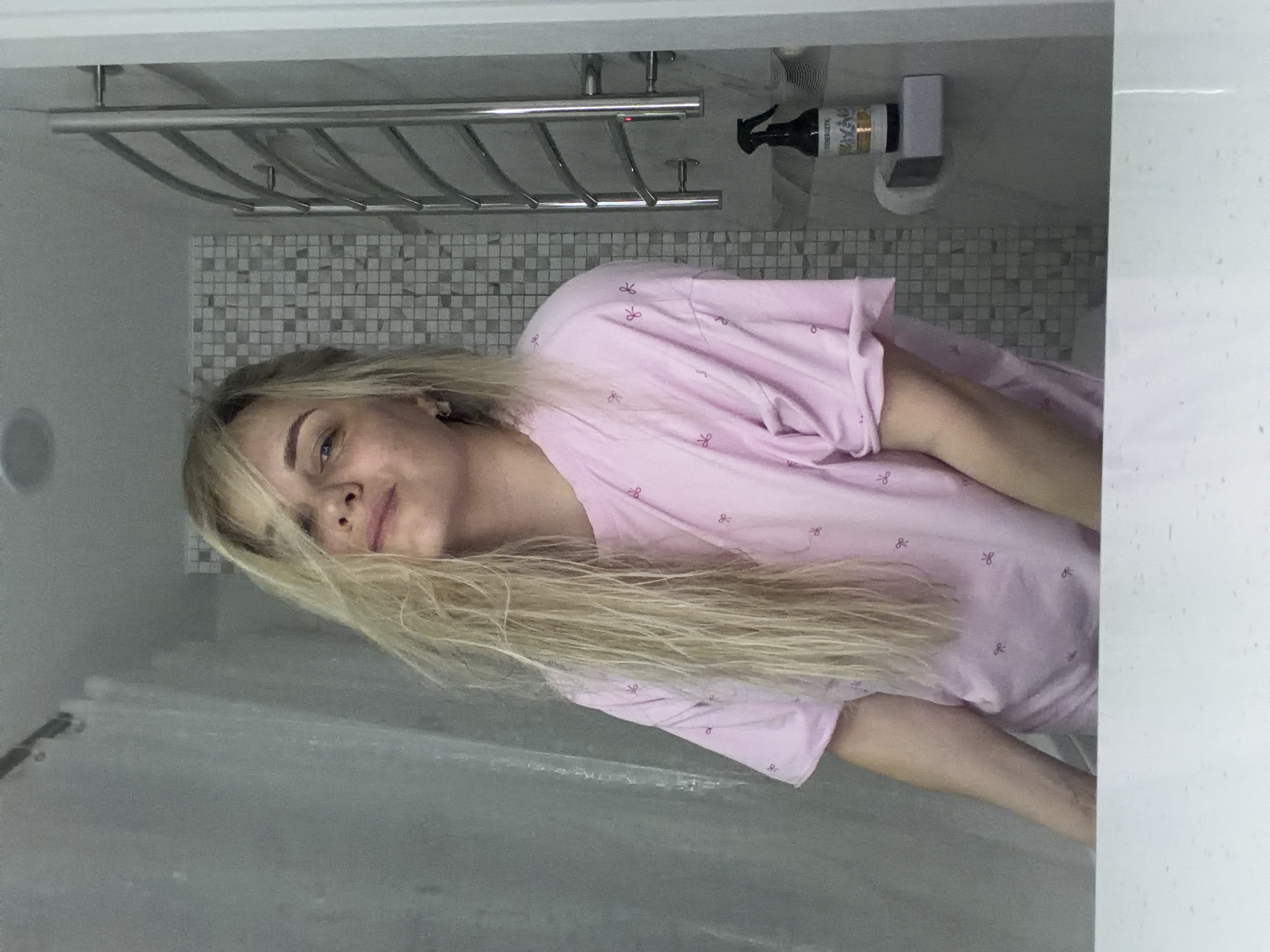 The image shows a blonde woman with light blue eyes and wearing a pink patterned top in what appears to be a bathroom. The background includes a tiled wall, a chrome towel rack, and a bottle of cleaning product. The foreground includes a shower curtain. It's impossible to determine whether the location is Votkinsk, Russia based solely on this image.