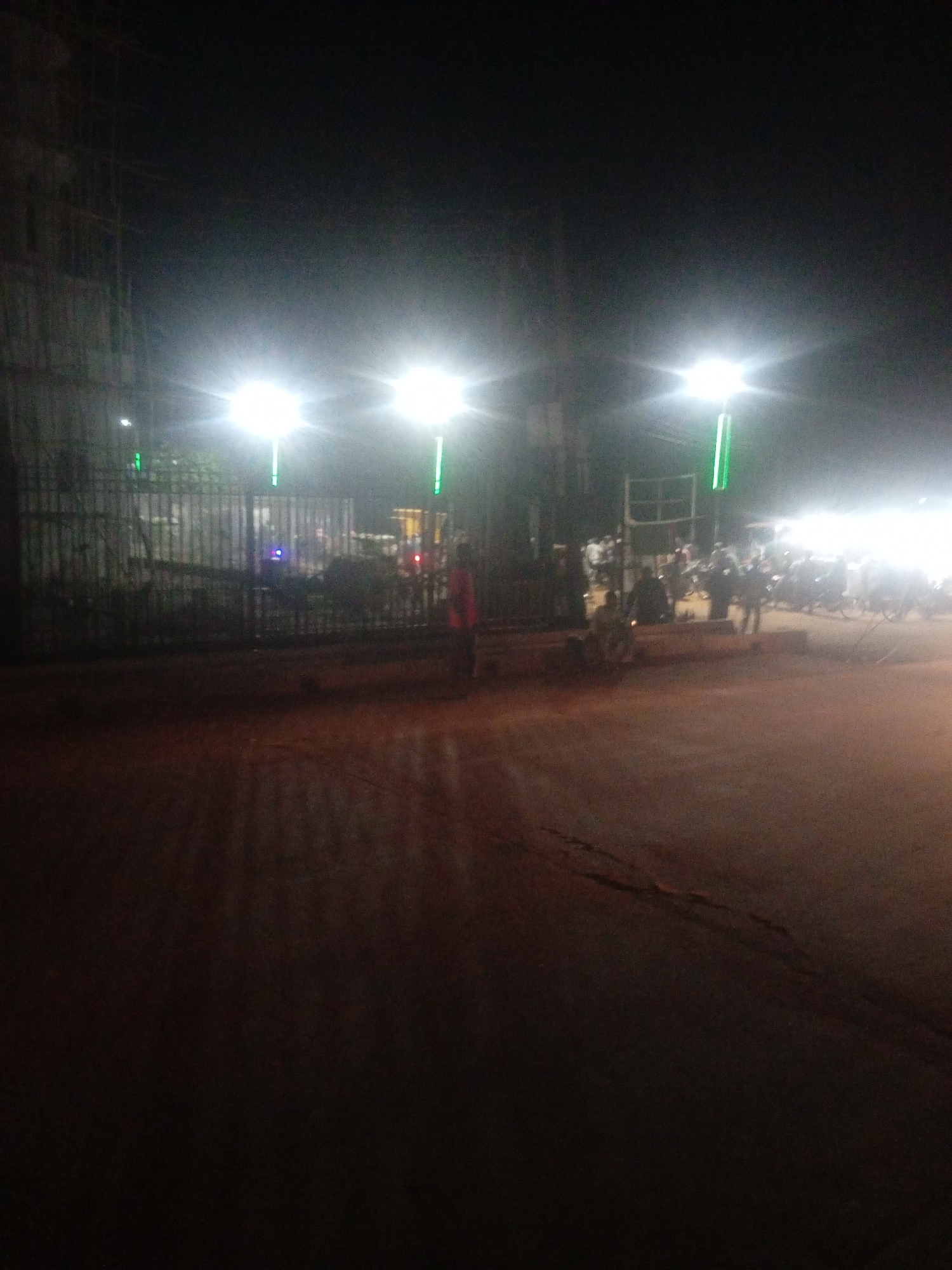 The image depicts an outdoor scene at night, illuminated by bright streetlights. The foreground is a dirt or gravel road, reflecting the light from above. In the midground, there is a dark metal gate with vertical bars. Beyond the gate, several people are visible, along with some parked motorcycles and what appear to be food stalls or temporary structures. The overall impression is of a bustling night market or gathering.
There are multiple bright lights, appearing to be street lamps or temporary lighting, casting a strong glow and creating starburst effects in the image. Two prominent lights are positioned on tall poles, one to the left and another slightly more to the right of the center. Smaller green lights are also visible, possibly decorative or part of the lighting setup. The sky is dark, indicating it is nighttime.
One person in the midground, to the right of the gate, is wearing a reddish-orange garment. Other figures are silhouettes against the bright lights, making their details indistinct. The atmosphere appears lively, suggested by the presence of many people and the activity implied by the stalls and parked vehicles. There is no visible text in the image. The setting suggests a public area, possibly a street or open space in a town or city, consistent with the provided location context of Tundun Wada South, Nigeria.