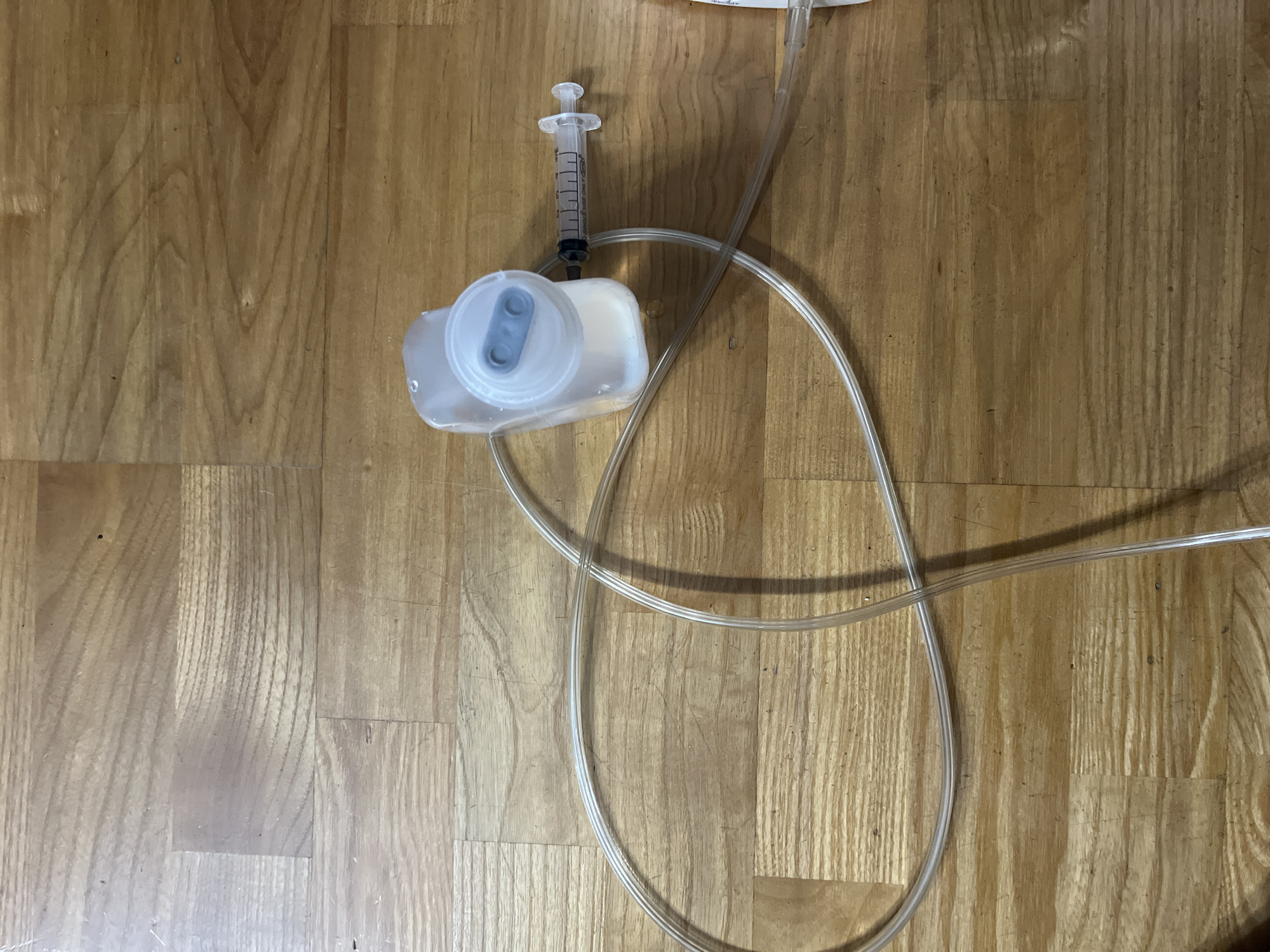 The image shows a clear plastic syringe attached to a clear plastic bag filled with a light-colored liquid. A clear plastic tube extends from the bag and forms a large loop before extending out of the frame. The items are placed on a wooden floor. The image is a close-up view and does not contain any information about the city of Saratov, Russia.