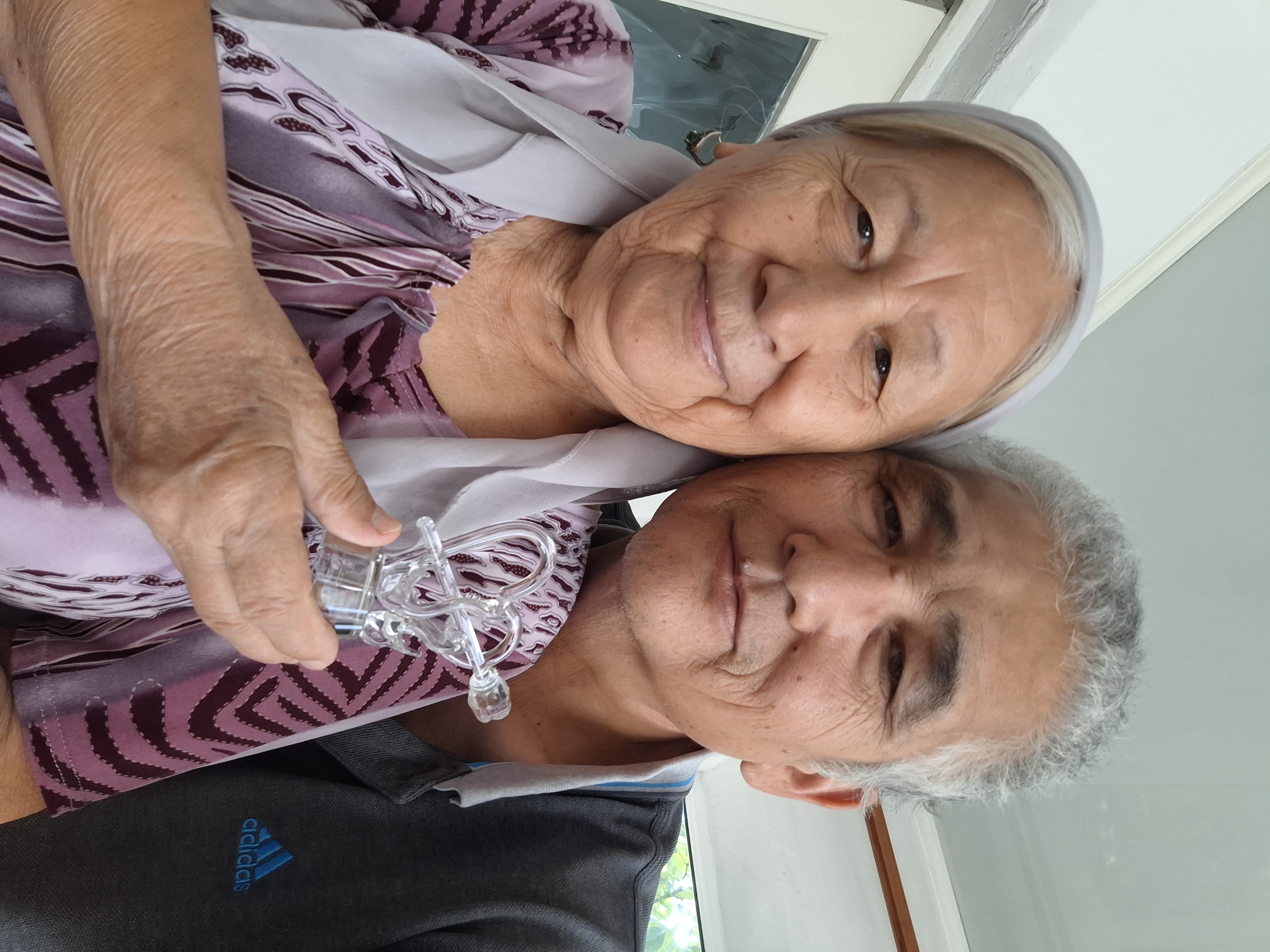 An elderly couple poses for a selfie, holding a small, clear glass object that appears to be a puzzle or a decorative piece. The woman on the left is wearing a light-colored headscarf and a purple patterned blouse. She has a gentle smile and is holding the glass object with her right hand. The man on the right has graying hair and is wearing a dark gray polo shirt with an Adidas logo. They are close together, with the man's arm around the woman. The background appears to be an indoor setting, possibly a home, with natural light coming from the left. The image was likely taken in Almaty, Kazakhstan, based on the context of the request.