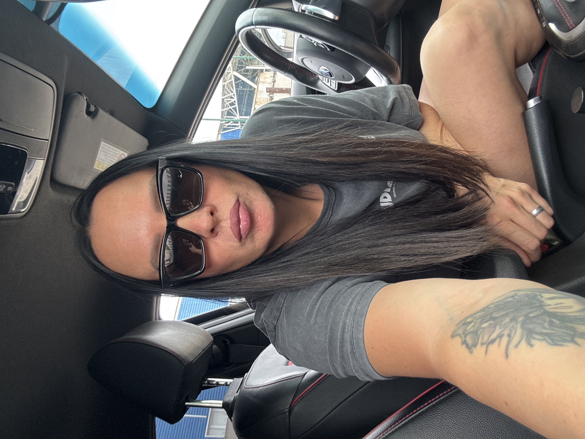A woman with long dark hair and sunglasses is taking a selfie inside a car. She is wearing a gray t-shirt and has a tattoo on her arm. A steering wheel is visible above her. Outside the car window, a scaffolding structure is partially visible with a blue building in the background. The exact location in Chaikovsky, Russia cannot be determined with the limited information provided in the image.