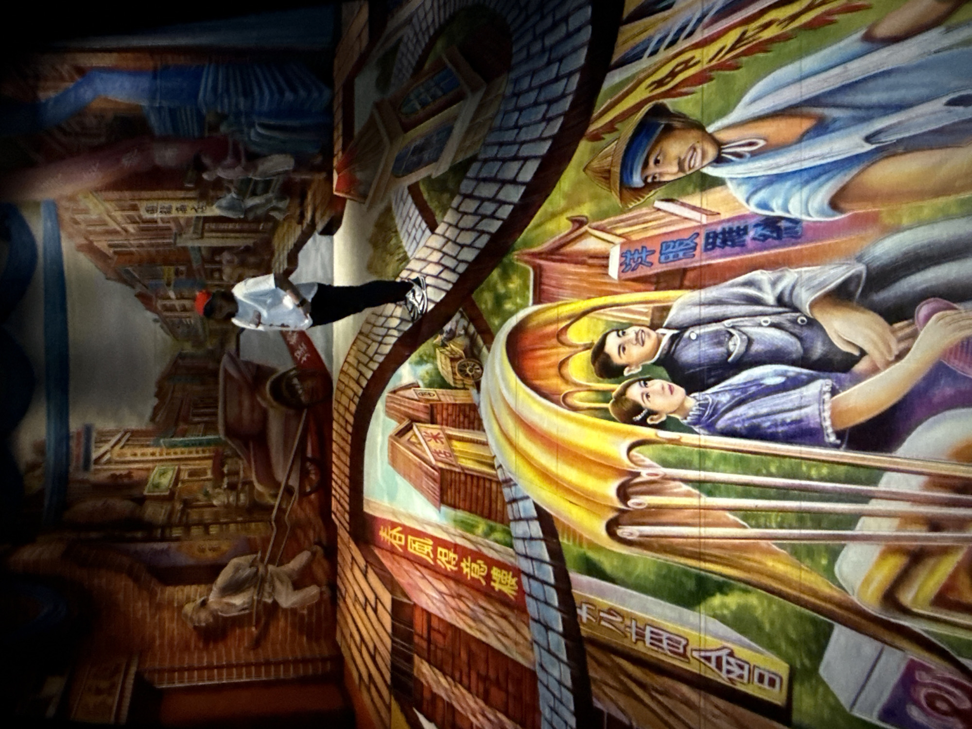 The image is a photograph of a mural painting that depicts a historical street scene in Taipei, Taiwan. The mural has a three-dimensional effect, making it appear as if the viewer is looking down a street.
In the foreground, three figures are prominently displayed. On the left, a woman in a purple traditional dress is seated, her hand resting on the arm of a man in a dark suit. To the right of the couple, a man wearing a straw hat and a blue garment is looking towards the viewer. Behind these figures, the mural continues with depictions of buildings with traditional Chinese architectural elements, colorful banners with Chinese characters, and what appears to be a covered walkway or bridge. The street scene also includes a rickshaw pulled by a man in the middle ground. Further back, more buildings and figures are painted, suggesting a bustling urban environment.
The overall impression is a nostalgic portrayal of life in Taipei during an earlier era. The lighting in the photograph highlights the painted details and creates a sense of depth. The perspective of the mural draws the viewer into the scene, as if they are walking through the depicted street.