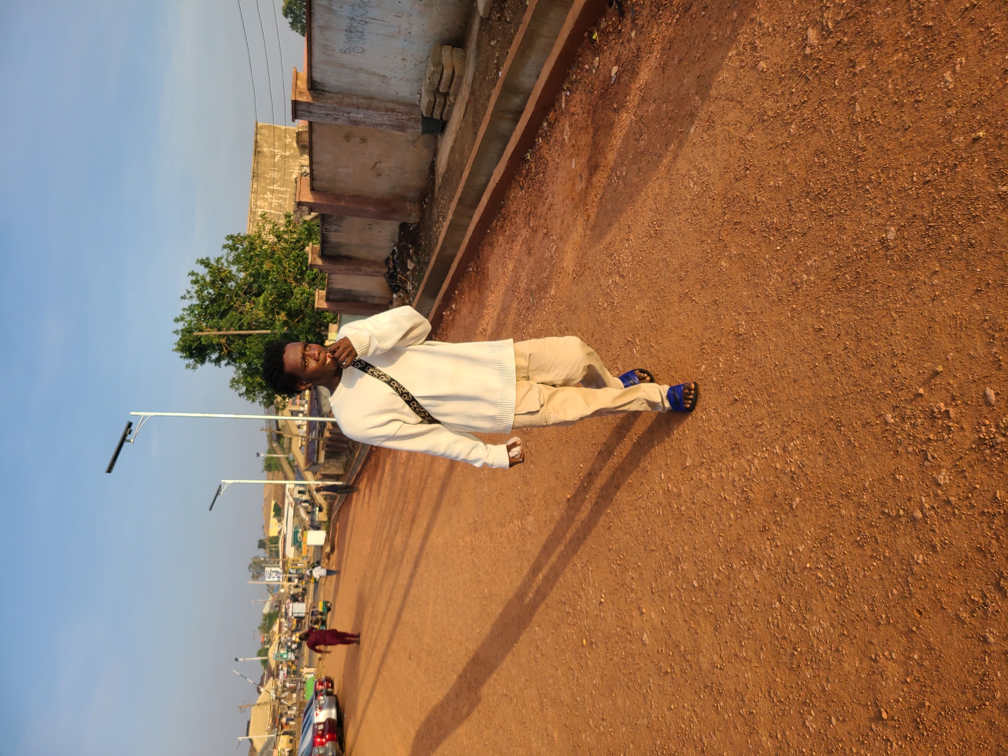 A long shot depicts a man lying diagonally across a dirt road in Jos, Nigeria. The road is a reddish-brown color and appears to be unpaved. The man is wearing a white sweater, tan pants, and blue sandals. He has a black strap or bag across his body. His shadow stretches across the road.
In the background, there's a view of the city, including buildings, streetlights, cars, and pedestrians. A concrete structure with some brickwork is visible closer to the man. The sky is blue.