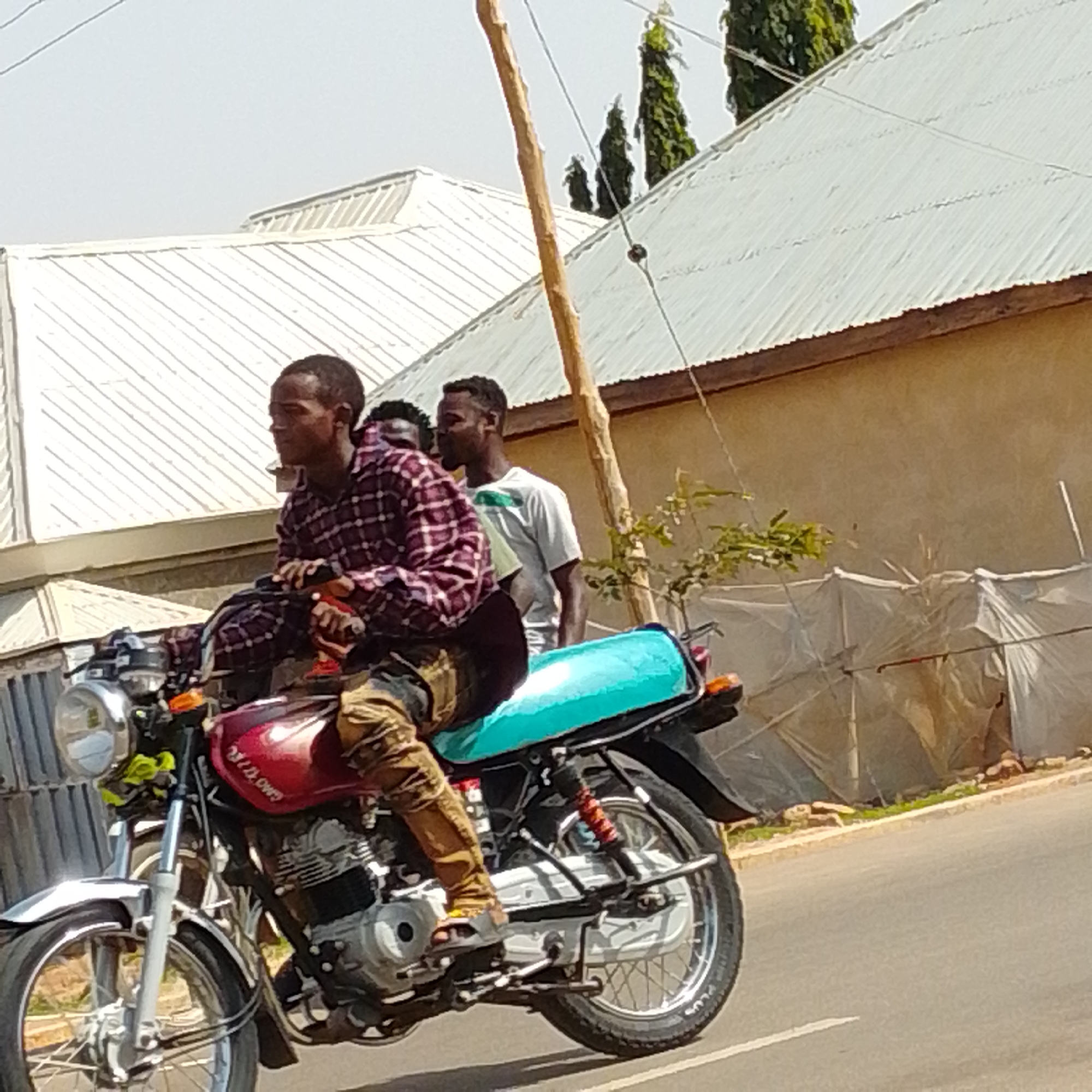 This is a medium shot, eye-level, outdoor photograph taken in the city of Gombe, Nigeria. The image captures three young Black men riding on a red motorcycle. The driver, wearing a red and white checkered shirt and brown pants, is on the left, looking forward. Behind him, a second man in a white t-shirt with green accents is seated in the middle, also looking forward. A third man, whose head is partially visible, is positioned behind the seated man. The motorcycle is moving on a paved road, with a brown building and a white corrugated metal roof structure in the background. A tall, thin wooden pole stands vertically to the right of the motorcycle, with some greenery and trees visible behind it. The sky is clear and bright. The motorcycle has a chrome headlight, a red fuel tank, and a turquoise seat. The men appear to be in motion.