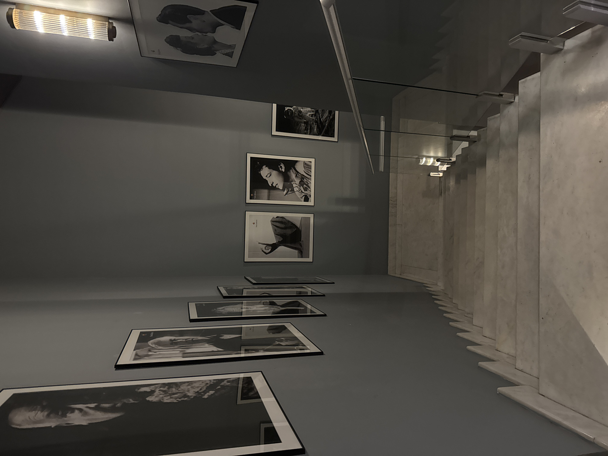 An interior view of a stairwell. Photographs are hung along a wall adjacent to white stairs with glass railing. A fluorescent light fixture is above the top photo on the wall. The location is Tbilisi, Georgia.