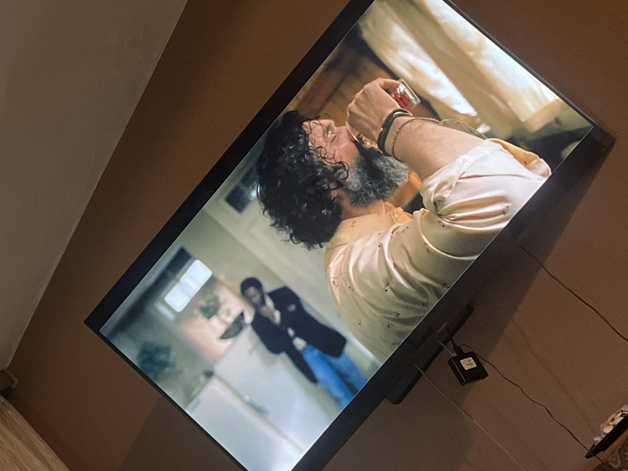 A tilted, medium-angle shot captures a flat-screen television displaying a scene from a film or TV show. The television screen is bright and dominates the frame, occupying the central and upper-left portion.
On the TV screen, the main subject is a man in the foreground, seen from the chest up, with a prominent salt-and-pepper beard and curly dark hair. He is wearing a light-colored, possibly cream or pale yellow, collared shirt and has several dark bracelets on his right wrist. He is actively drinking from a small, clear glass with red contents, appearing to be taking a shot. His head is tilted back, and his eyes are closed or squinted, conveying an intense or perhaps slightly pained expression as he drinks.
In the blurred background of the TV screen, another man is visible standing in what appears to be a doorway or hall. He is wearing a dark jacket and blue jeans. The overall setting within the TV show seems to be an indoor environment, possibly a home, with hints of curtains in the brighter background behind the drinking man and indistinct domestic elements behind the second man.
Outside the television frame, the surrounding environment is a light brown wall or furniture surface. The TV is positioned at an angle relative to the viewer. To the bottom right of the TV, a small black electronic device, possibly a power adapter or converter, is visible with wires extending from it. Text on this device is present but unreadable due to blurriness. The lighting in the room where the photo was taken is ambient, with the TV screen itself being the primary light source for the depicted scene. The time of day is not discernible. The location context is Yardzhilovtsi, Bulgaria.