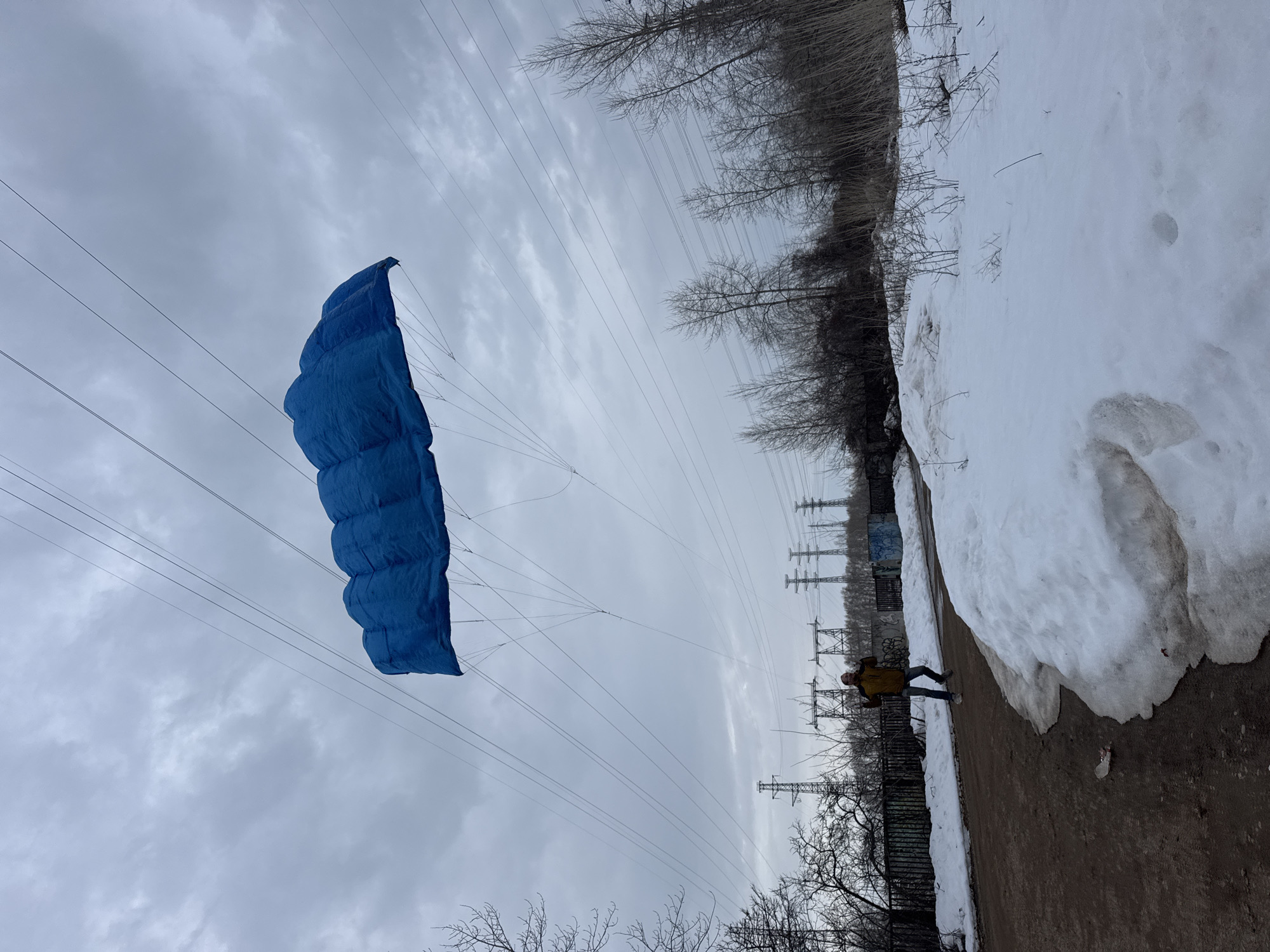 A low-angle shot shows a blue parachute suspended in the air near power lines in a snowy, urban landscape. A person in a yellow jacket stands on a path adjacent to a snowdrift and a fence. Bare trees are present on the horizon with more power lines and towers behind them. The sky is overcast. The location is speculated to be in the city of Chaikovsky, Russia.