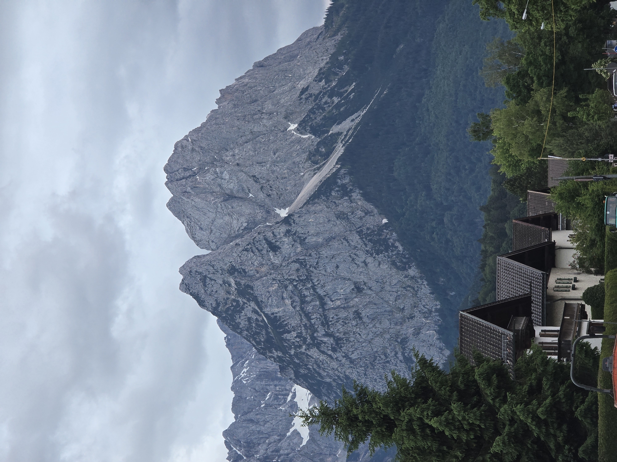 This image captures a landscape in Garmisch-Partenkirchen, Germany, under an overcast sky. The prominent feature is a vast, rugged mountain range, likely a part of the Wetterstein massif, dominating the background. These mountains are characterized by their grey, exposed rock faces with visible stratification and sparse patches of snow in higher elevations and shaded gullies. The lower slopes of the mountains are densely covered with dark green coniferous forests, which transition into the rocky peaks.
In the mid-ground and foreground, a residential area is visible. On the left, several traditional Bavarian-style houses with dark brown gabled roofs, light-colored facades, and wooden balconies and shutters are partially obscured by a large, dark green coniferous tree. Further to the right, other deciduous trees with lighter green foliage are present. The very bottom foreground hints at a street or public area, showing parts of what appears to be a red/orange piece of machinery (possibly a small vehicle or construction equipment), green hedges, and faint glimpses of street signs and lampposts. No individuals are discernible within the frame. The overall impression is of a mountainous town environment under a cloudy day.