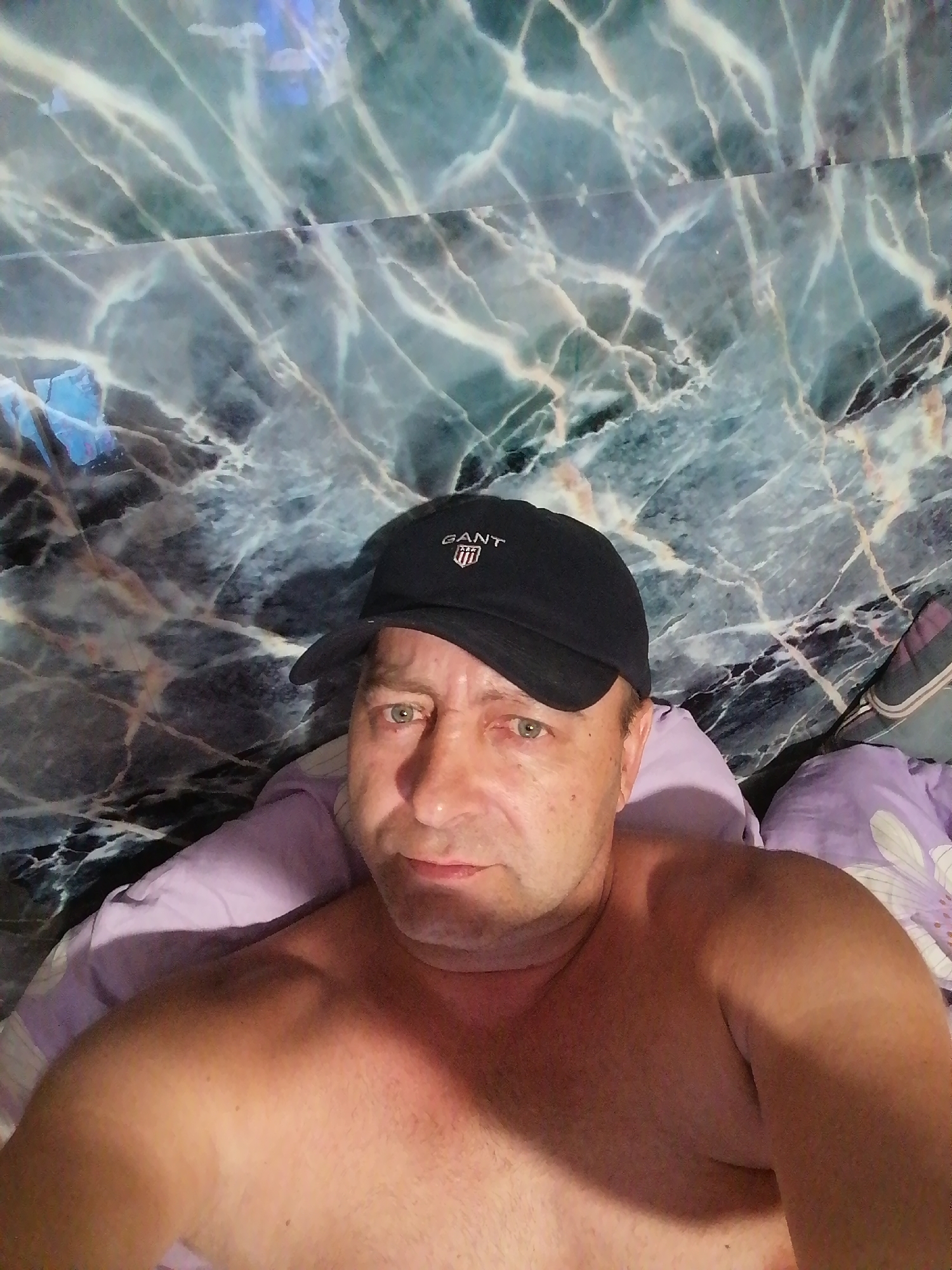 The image shows a close-up, selfie-style shot of a shirtless man wearing a black baseball cap with the "GANT" logo. He is lying down, with his head and upper torso filling the frame. The background appears to be a wall with a marble-like pattern in shades of green and blue. There are pillows visible around the man, one of which is purple with a floral design. The lighting is somewhat dim, creating shadows on the man's skin. No specific location in Novopokrovka, Russia is discernible from the image.