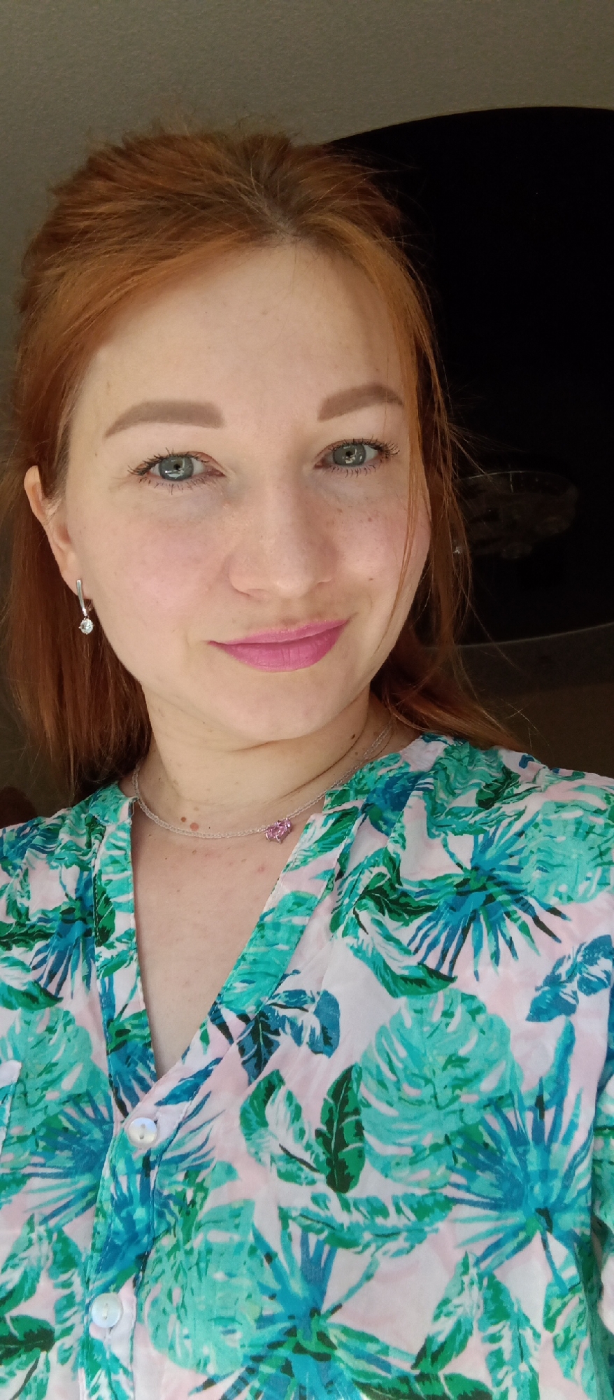 A close-up shot shows a fair-skinned woman with red hair and blue-green eyes. She is wearing light pink lipstick and a patterned shirt with tropical leaf motifs in shades of green, white, and blue. She is also wearing a silver chain necklace with a pink pendant and silver earrings with a small, clear stone. The background is dark and indistinct. Location: Unknown, Unknown.