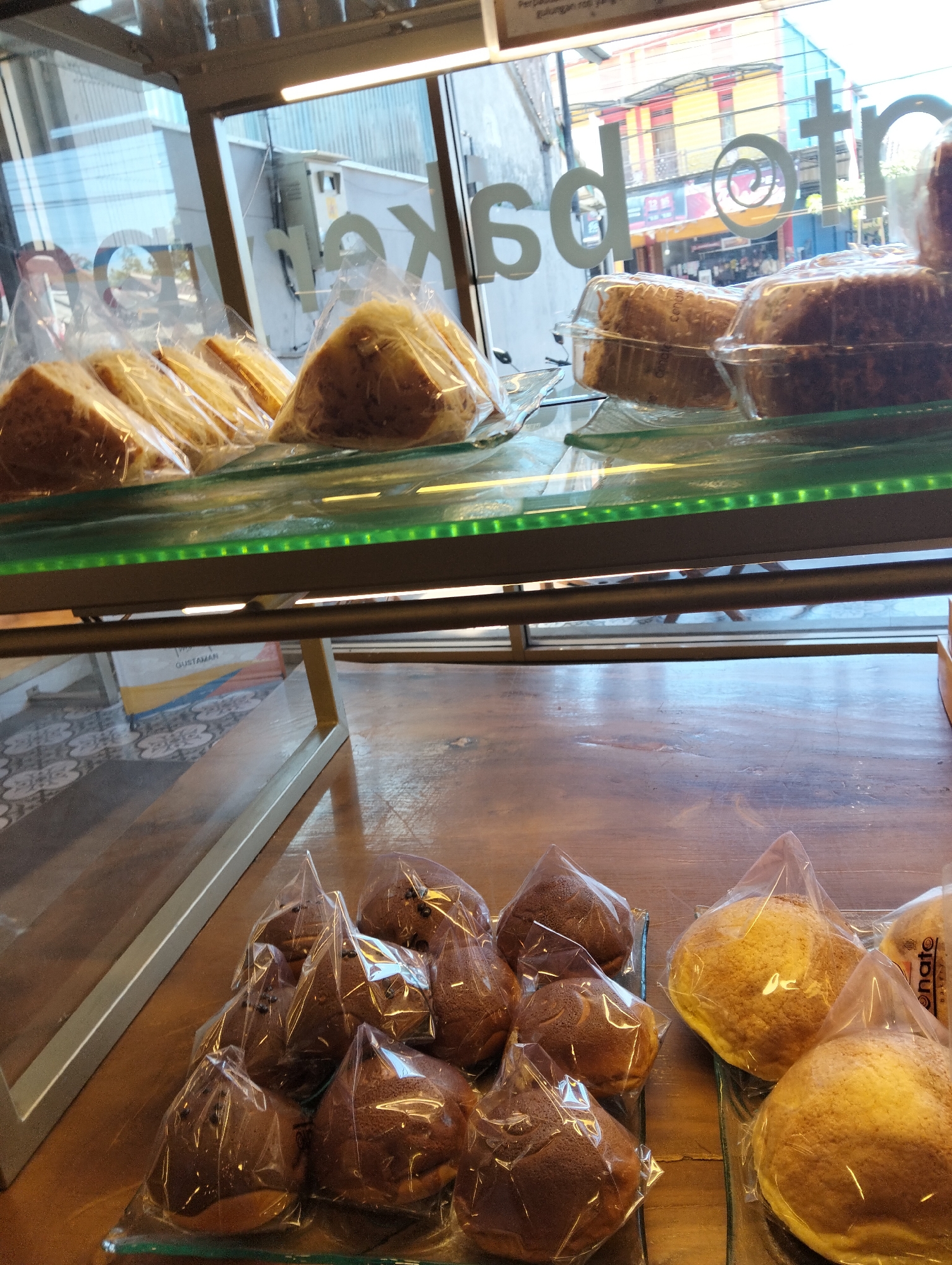 In a bakery display case in Sumberberas, Indonesia, pastries are arranged on glass shelves. The top shelf holds sliced cakes wrapped in plastic, with slices of bread visible behind them. The lower shelf showcases an assortment of baked goods: several individually wrapped chocolate-colored buns with visible chocolate chips, and two larger, golden-brown, round pastries in plastic wrap. The bakery has large windows that look out onto a street with colorful buildings. A sign with white lettering, possibly the bakery's name, is visible on the window. The interior of the display case is illuminated by green and white LED lights. The scene is captured from a slightly elevated perspective, looking down into the display.
