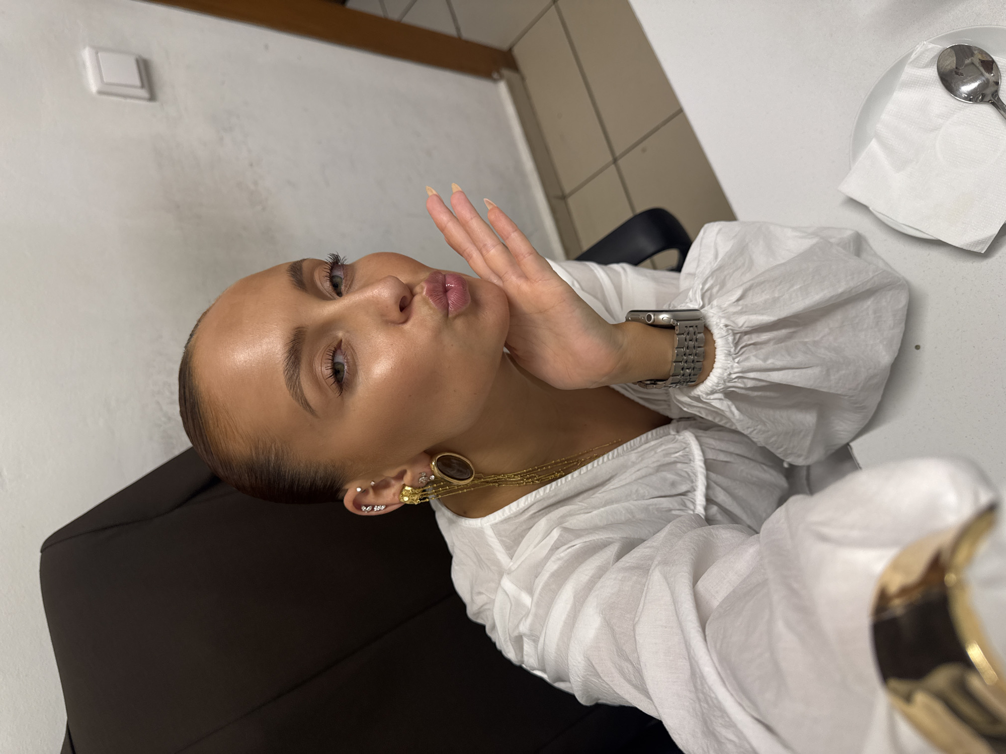 A woman with blonde hair pulled back and wearing a white shirt is seated at a desk, leaning forward and blowing a kiss towards the camera. She has large earrings in her ears and is wearing a watch on her left wrist. The background shows a black chair, a white wall with a light switch, and a tiled floor. There is a white plate with a napkin and spoon on the desk in the foreground. The scene is indoors and appears to be an office or a similar setting.