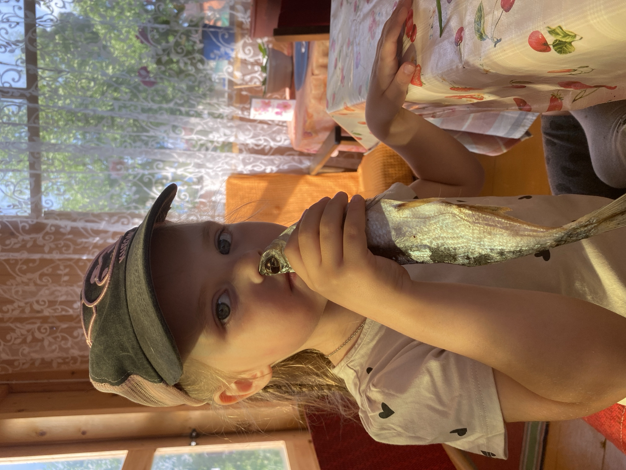 A young girl wearing a dark baseball cap and a white t-shirt with a heart pattern holds a dried fish up to her nose. She is seated at a table covered with a light-colored tablecloth featuring a pattern of cherries and strawberries. Behind her, a window with lace curtains reveals a sunlit outdoor scene with trees. The environment suggests a casual, possibly rural, setting, perhaps a dacha or summer house in Votkinsk, Russia.