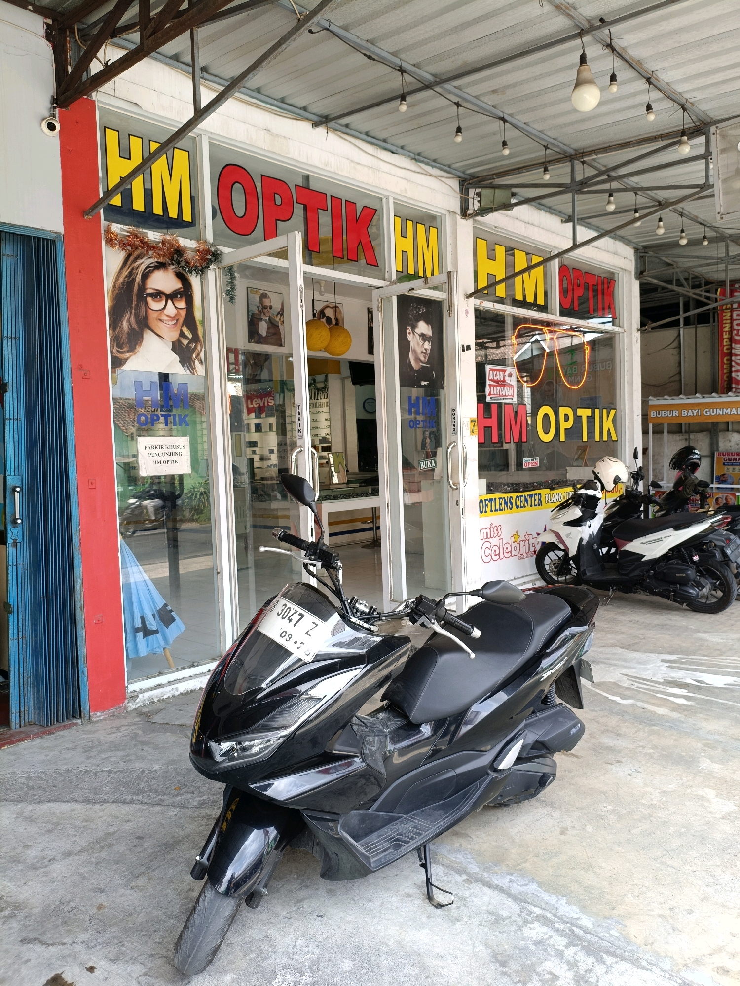 The image captures the storefront of "HM OPTIK" in Maguwoharjo, Indonesia, a business specializing in optical goods. The shop's facade features large glass windows with prominent yellow and red lettering spelling out the business name. Inside, eyewear displays are visible, along with advertisements and posters, including one featuring a woman with glasses and another of a man in glasses. A neon sign shaped like glasses is also displayed in the window.
In front of the shop, parked on a concrete pavement, are several motorcycles. The most prominent is a black scooter in the foreground, with a license plate that reads "B 3047 Z 09-19". To the right of this, two other motorcycles are parked, one white and maroon, and another black. The surrounding environment suggests an urban or commercial street setting, with a corrugated metal roof supported by metal beams overhead, and hanging lightbulbs. A blue folding door is visible on the left side of the frame, and a red pillar stands next to it. The overall scene is well-lit, indicating it was likely taken during daylight hours.