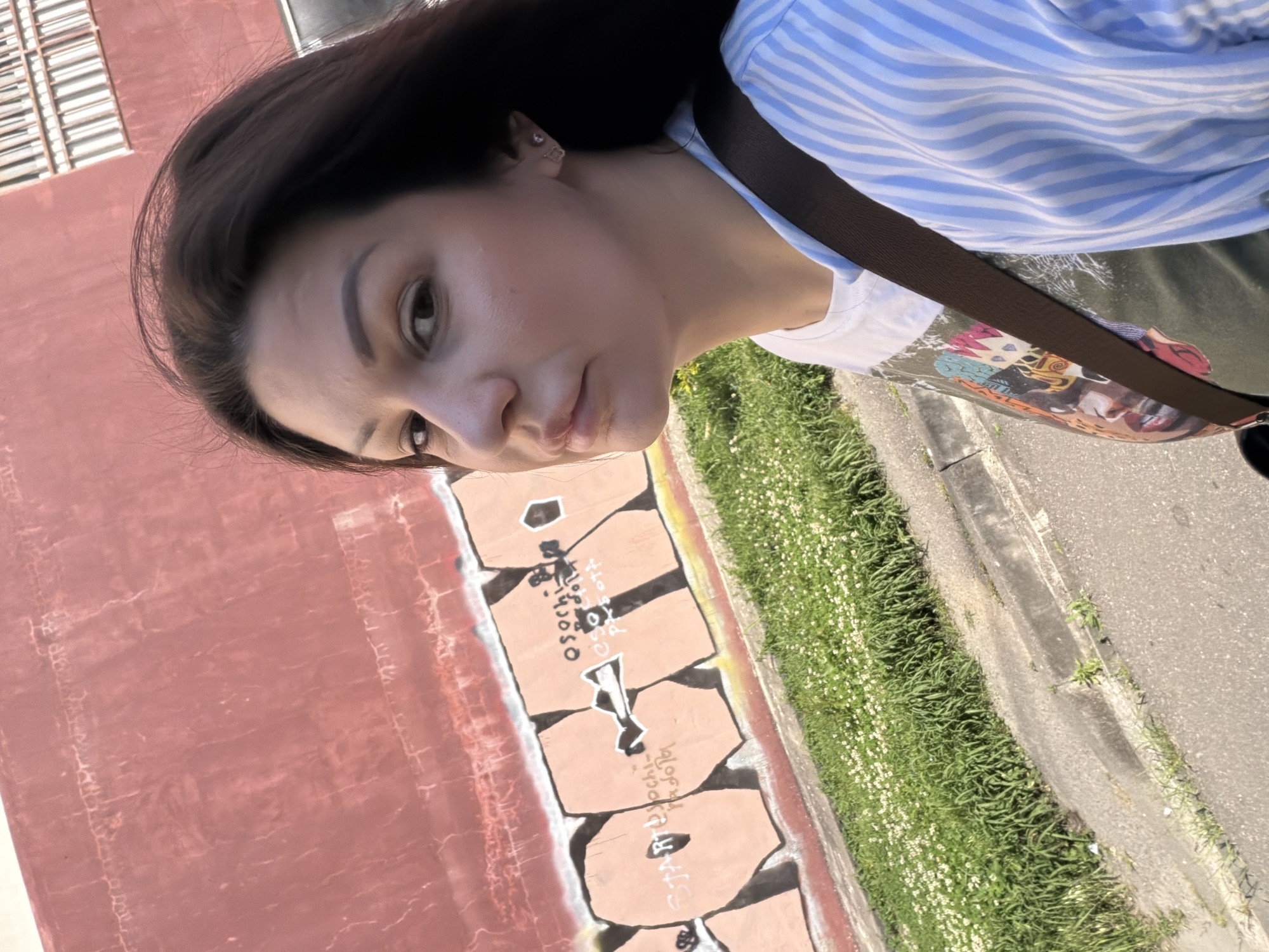 An adult female is captured in a self-portrait or close-up photograph outdoors during daylight hours. She has dark brown hair and a fair complexion, looking directly at the camera with a neutral expression. She wears small earrings and is dressed in multiple layers, including a light blue and white striped long-sleeved top, a dark olive green garment, and a lighter t-shirt with a graphic design. A dark brown strap, likely from a bag, crosses her torso.
Behind the subject is a prominent, weathered red-orange wall exhibiting areas of faded paint and cracking. Along the lower section of this wall, block-style graffiti in a lighter peach color with dark outlines is visible. Overlaid on this graffiti are lighter inscriptions in Cyrillic, prominently featuring variations of "Osochi" or "Sochi," along with words like "START," "radota," "esodn," and "pxsott," confirming the location in Sochi, Russia.
Below the wall, a strip of green grass populated with small white flowers borders a concrete curb, which separates it from an asphalt road. In the upper right background, a section of a building with multiple horizontal barred windows or grates is partially visible, indicating a larger structure beyond the wall.