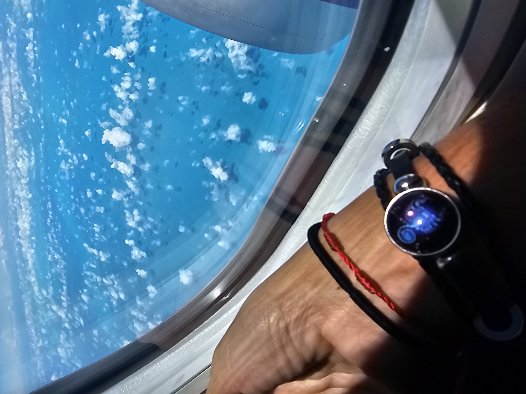 The image shows a view from an airplane window, overlooking a turquoise ocean with scattered clouds. A person's hand, adorned with bracelets (one with a watch face displaying a constellation), is visible in the foreground. The location is likely over the Bahamas, based on the ocean color, but the specific city is unknown.