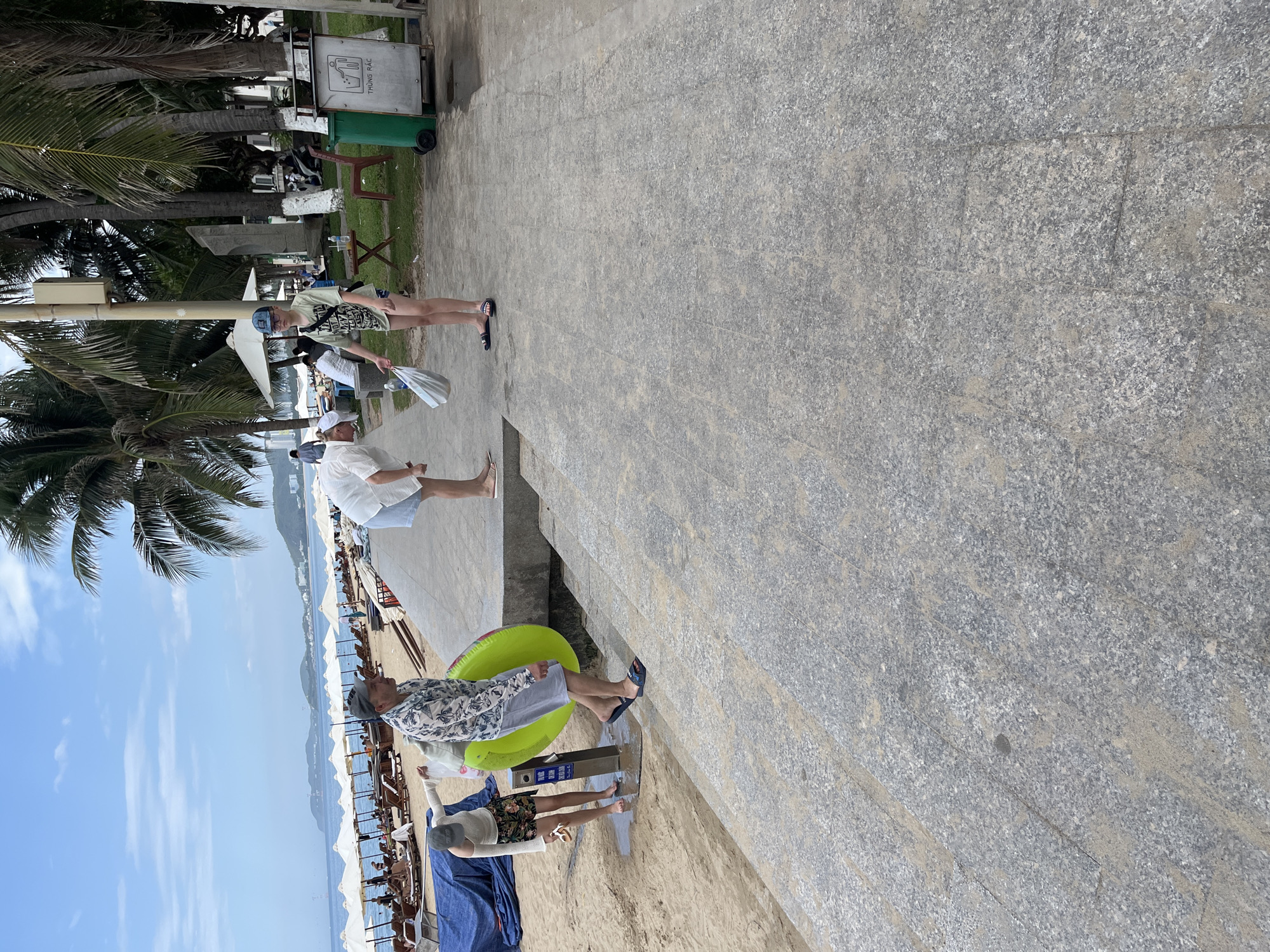 In Nha Trang, Vietnam, a beach scene unfolds with a group of people near the shoreline. A man in a patterned shirt and grey shorts carries a bright green inflatable ring. Beside him, a woman in a white top and floral shorts bends down. Another person, a young individual in a light green t-shirt and dark shorts, stands with a plastic bag. Further down the beach, rows of white umbrellas shade lounge chairs, and palm trees line the area. The ground is a mix of sand and paved pathways, with a green trash bin visible in the background. The sky is clear blue with some scattered clouds.