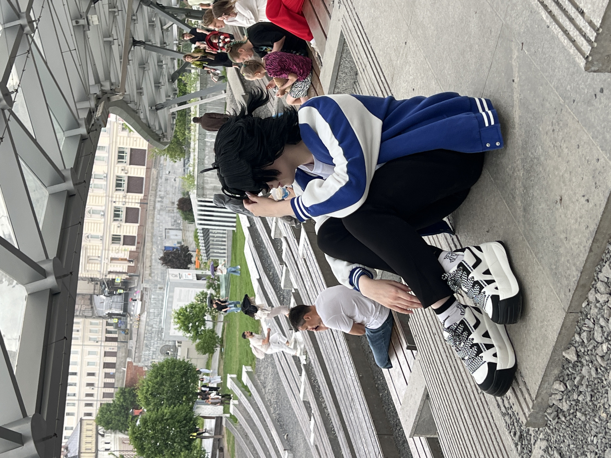 A ground-level, medium shot shows a person with black hair and wearing a blue and white jacket, black leggings, and black and white platform sneakers sitting on a grey bench. The individual appears to be using a mobile phone. The image also shows many other people sitting on adjacent benches. Beyond the benches, there is a green space, a street, and building in the background. The location is Zaryadye Park in Moscow, Russia.