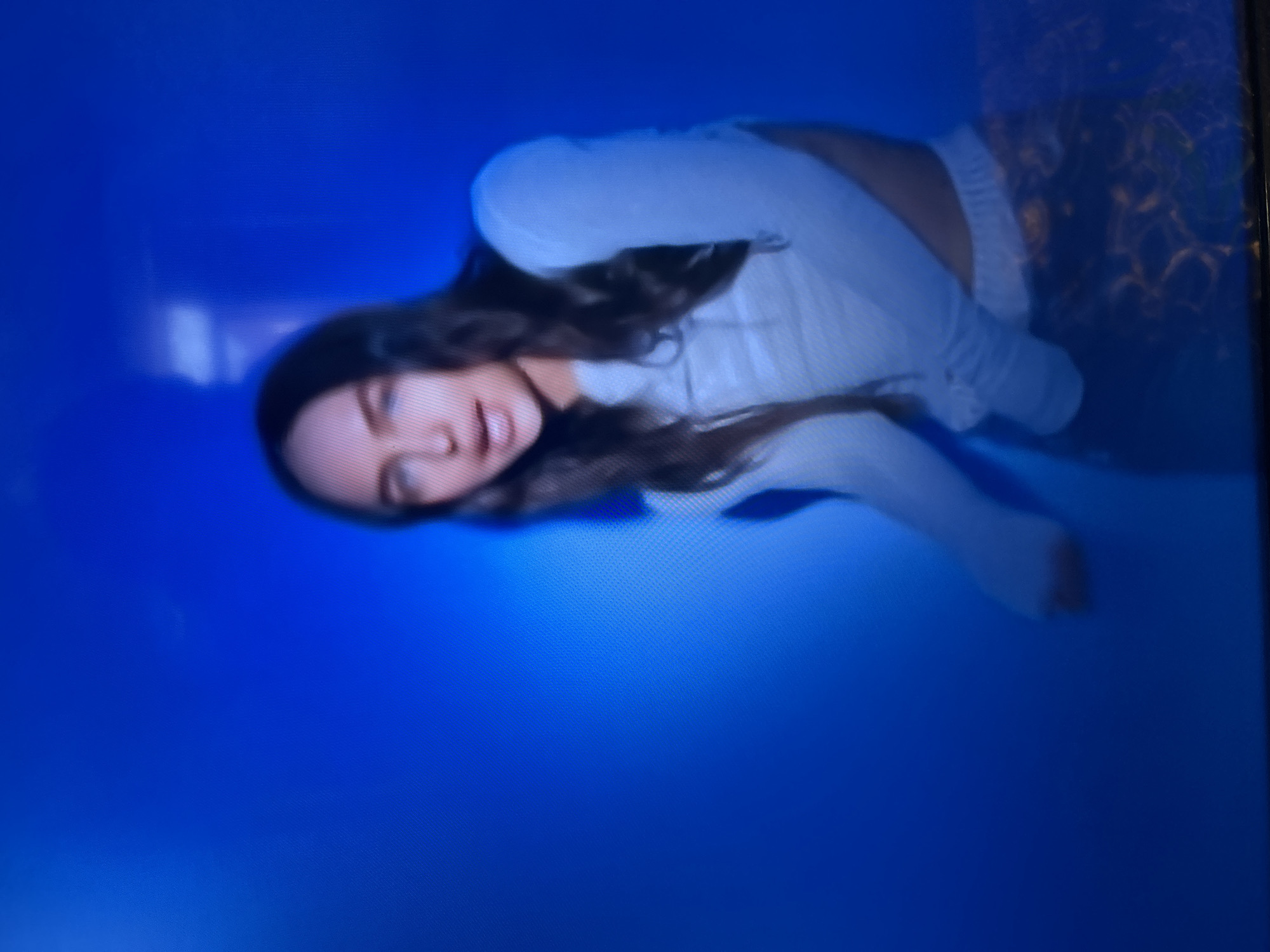 The image shows a blurry, medium shot of a young woman with long, dark hair and fair skin. She is wearing a white long-sleeved top and patterned pants. She is looking forward and her mouth is slightly open. The background is a solid, bright blue with some lighter blue and white highlights, suggesting a studio setting with dramatic lighting. There is no discernible information in the image that could indicate a specific location in Gelendzhik, Russia, or any other city.