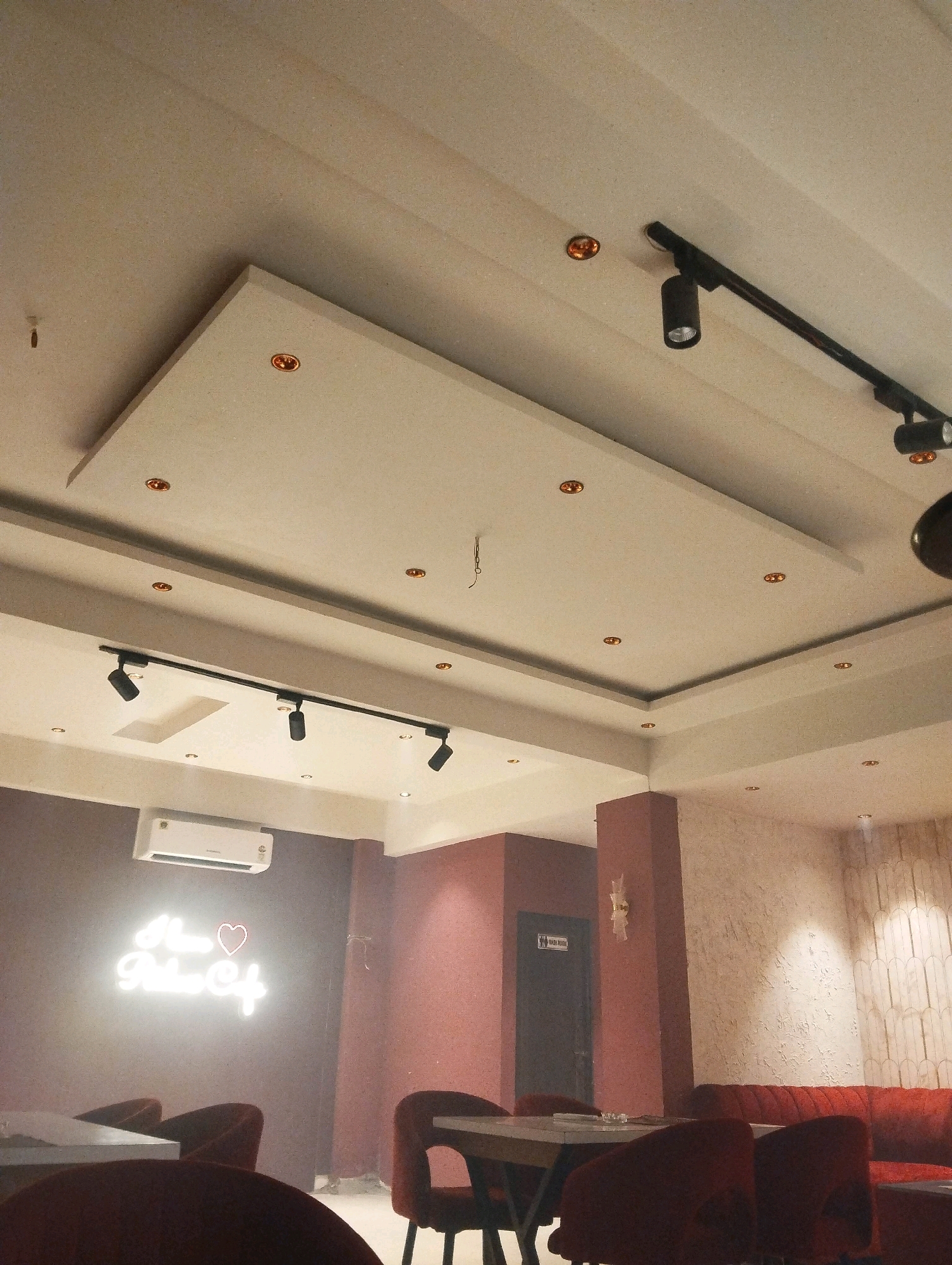 The image shows the interior of a cafe, likely in Lucknow, India, judging by the neon sign on the wall that reads "I Love Robin Cafe". The cafe appears to be dimly lit, with ambient lighting coming from recessed ceiling lights, track lighting, and the neon sign itself. The ceiling is a multi-layered design with recessed areas and exposed beams. The walls are painted in muted colors, with a textured finish on the far right wall.
In the foreground, there are several plush, dark red chairs and a table. The chairs have a velvet-like texture and a classic design. In the mid-ground, a door marked "Wash Room" is visible, with a decorative sconce above it. Further back, a long red sofa is visible, hinting at more seating arrangements. An air conditioning unit is mounted on the wall above the neon sign. The overall impression is of a cozy and somewhat dimly lit cafe, suitable for a relaxed atmosphere.