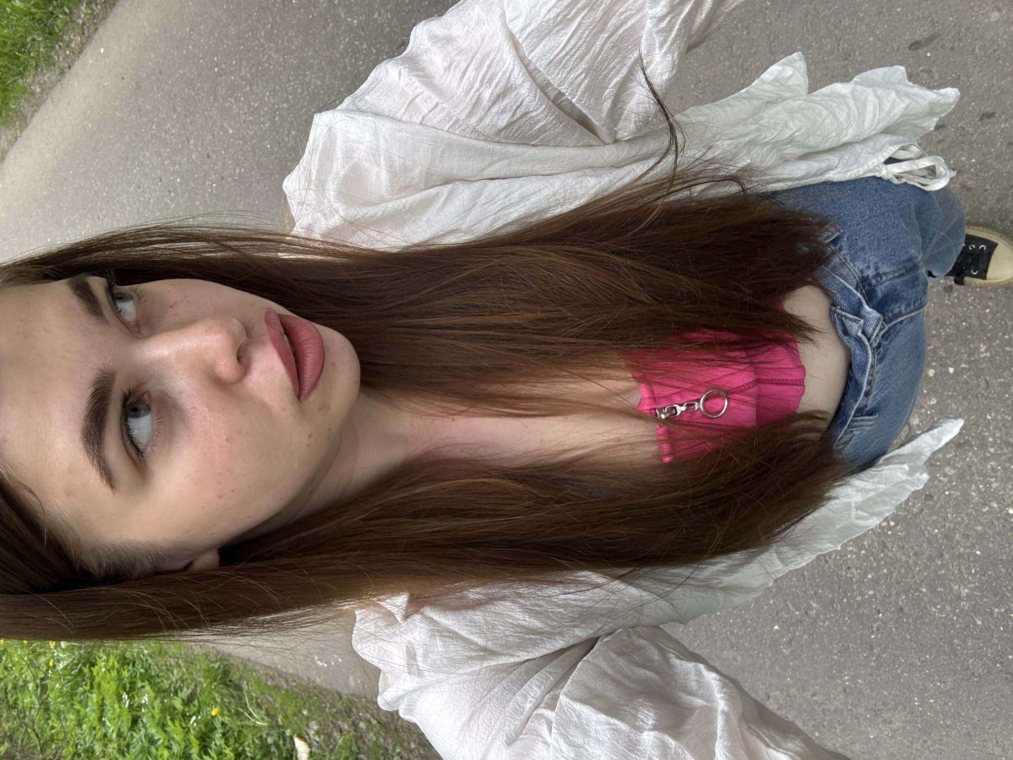 A young woman is looking up at the camera. She has brown hair, fair skin, and is wearing a pink top under a white blouse, blue jeans and white shoes with black stripes. The background consists of gray pavement and grass. The location is not specified, and the prompt does not instruct me to locate the city of Meshchery.
