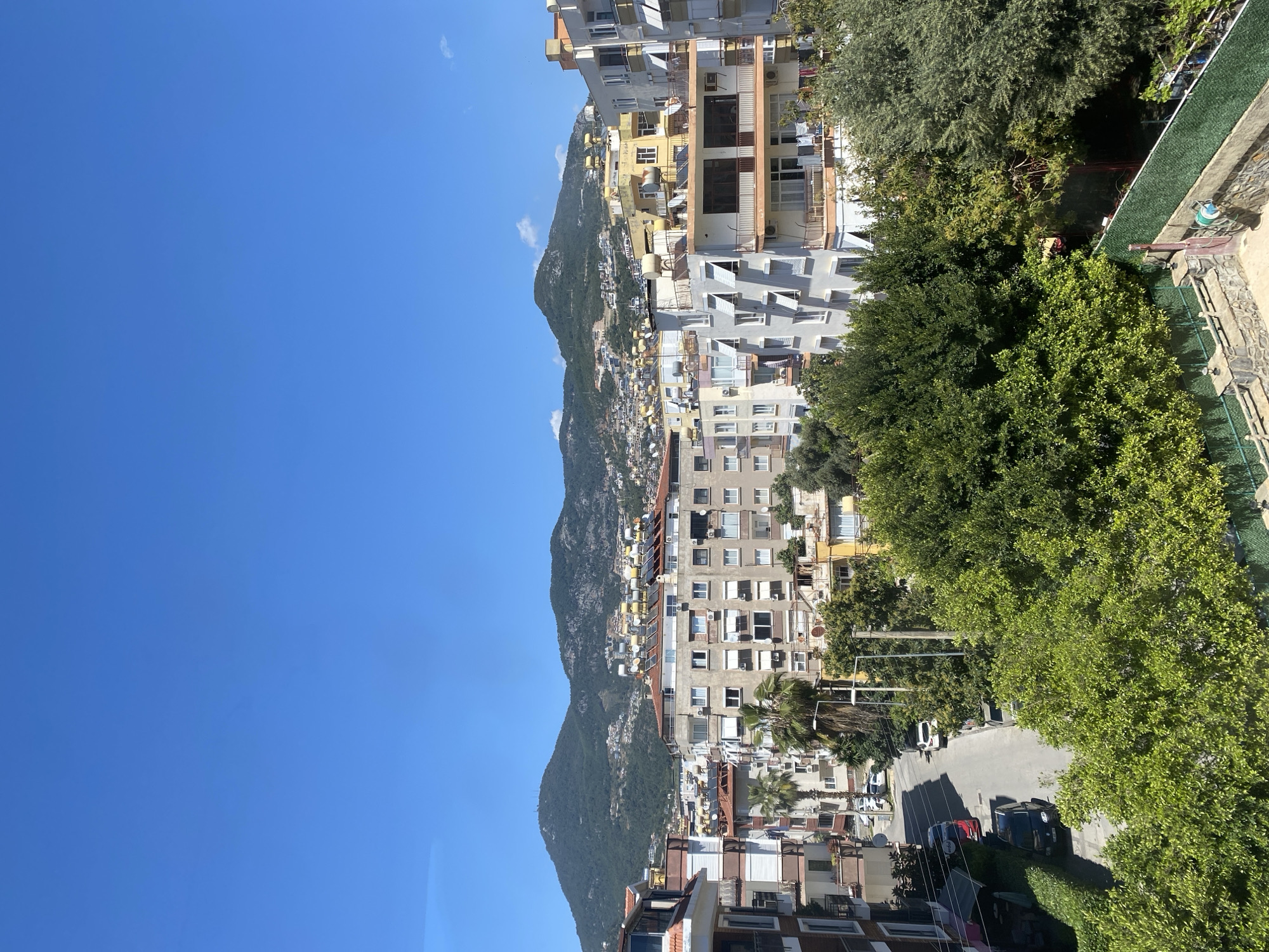 The image depicts a cityscape in Alanya, Turkey, positioned against a backdrop of a mountain range under a clear blue sky. The view is slightly elevated, possibly from a building. The mountain range is green with vegetation. The city itself consists of diverse buildings, ranging from low-rise residential to taller apartment complexes. A street is visible, populated with parked cars and streetlights, fringed with trees and vegetation. The angle of the photograph might suggest a location along or near a slope or hillside within Alanya.