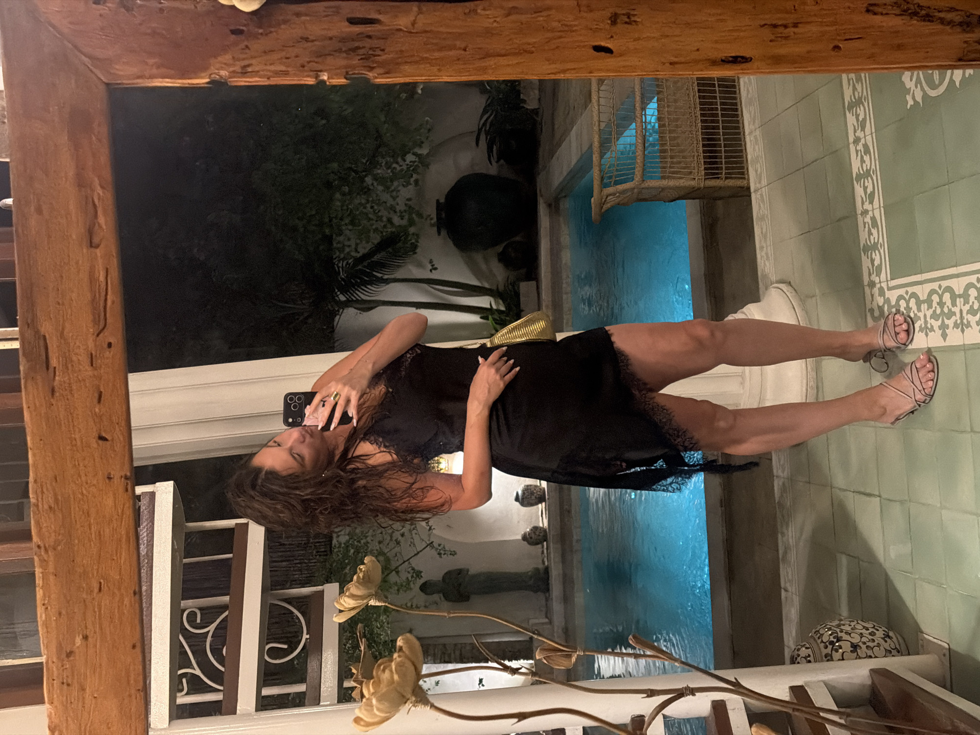 The image shows a woman taking a mirror selfie at night. She is wearing a black lace-trimmed slip dress and strappy sandals. The setting appears to be an outdoor area with a swimming pool and lush greenery. In the reflection, the woman is holding up a smartphone to take the picture. A large, rustic wooden beam frames the top of the image. The pool is lit with blue light, indicating it is nighttime. The presence of tropical plants and the vibrant blue of the pool suggest a warm climate, consistent with the North Kuta, Indonesia location context. The woman's pose and the setting evoke a sense of relaxation and enjoyment.