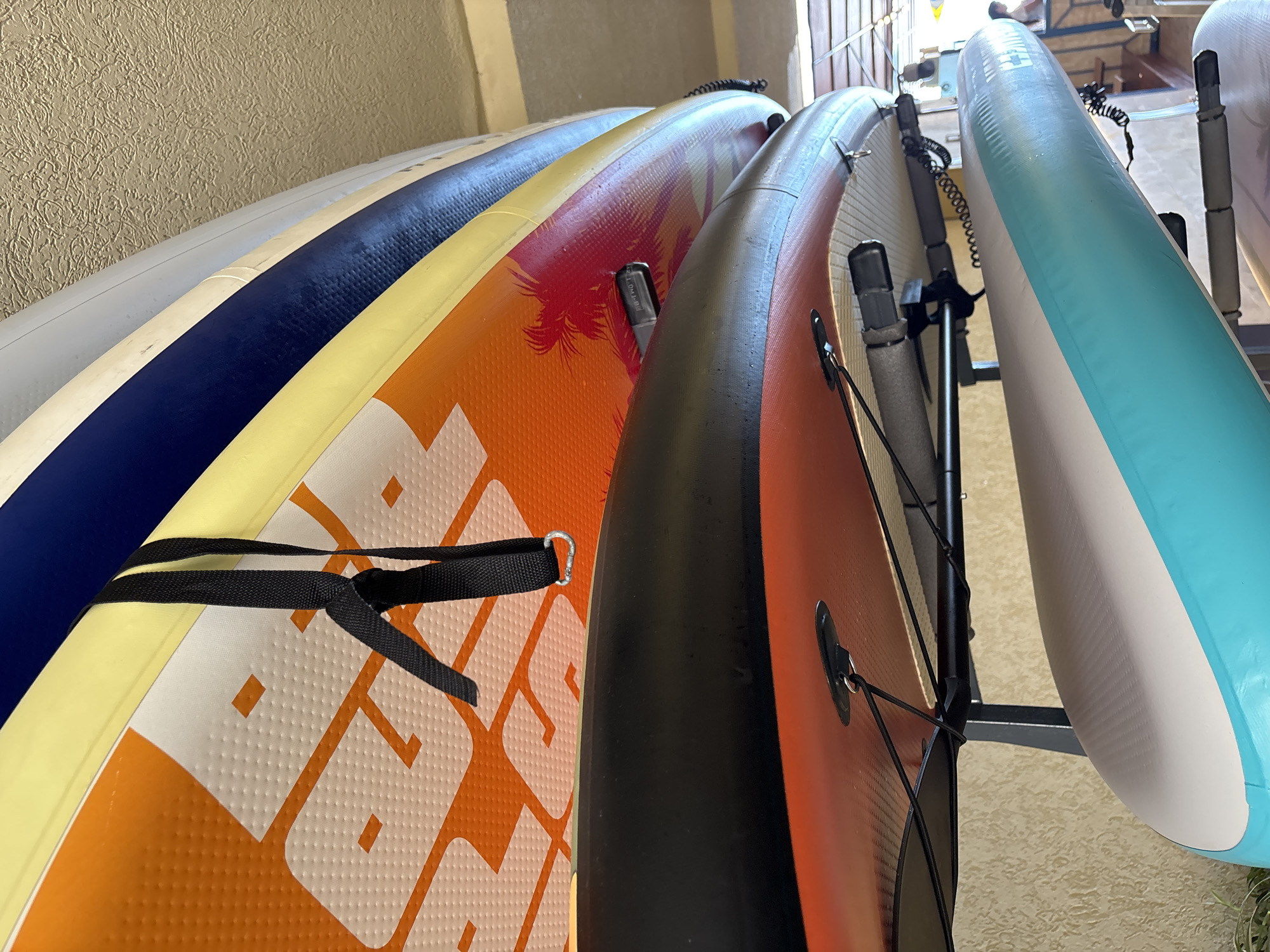 An indoor storage rack holds several paddleboards, stacked vertically. The top paddleboard is orange with a palm tree graphic and a black strap with a metal ring. Below it is a black paddleboard, followed by a red and white paddleboard. The bottom paddleboard is white with a turquoise stripe. The paddleboards are positioned in what appears to be a storage area or garage, with a textured ceiling and wall. There are no people visible in the image. The overall scene is indoors, suggesting a climate-controlled environment for storing watercraft.