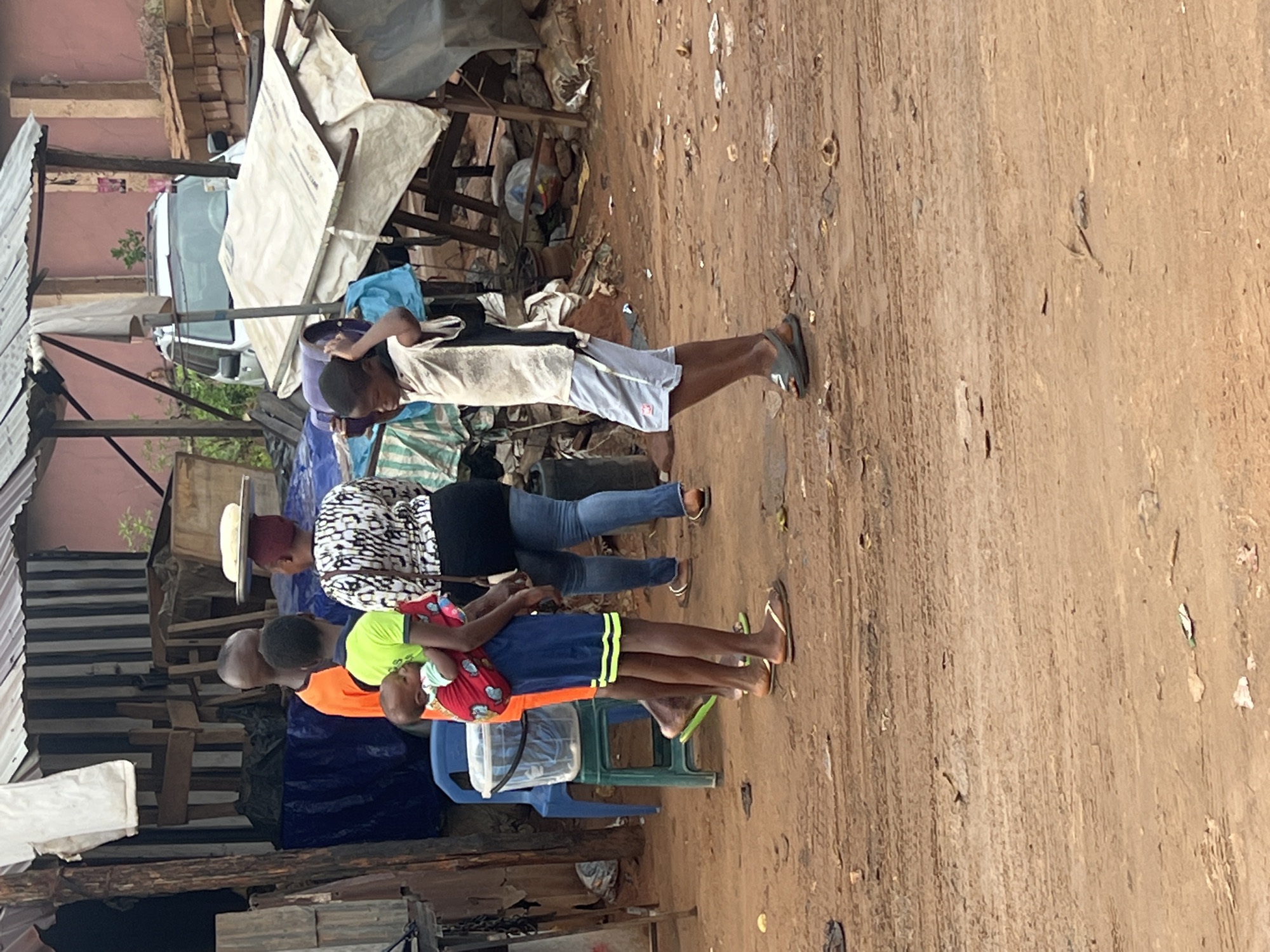 The image depicts a scene in Onitsha, Nigeria. A group of individuals, including children, is present in what appears to be a market or residential area with makeshift structures. A woman with a white hat is standing next to a child being held in an orange shirt in front of blue plastic chairs. Another child is standing with a gray shirt and blue shorts. The ground is a mixture of bare earth and some pavement. Debris and various objects are scattered around. The structures and tents are constructed out of metal poles, white tarp, and scrap wood.