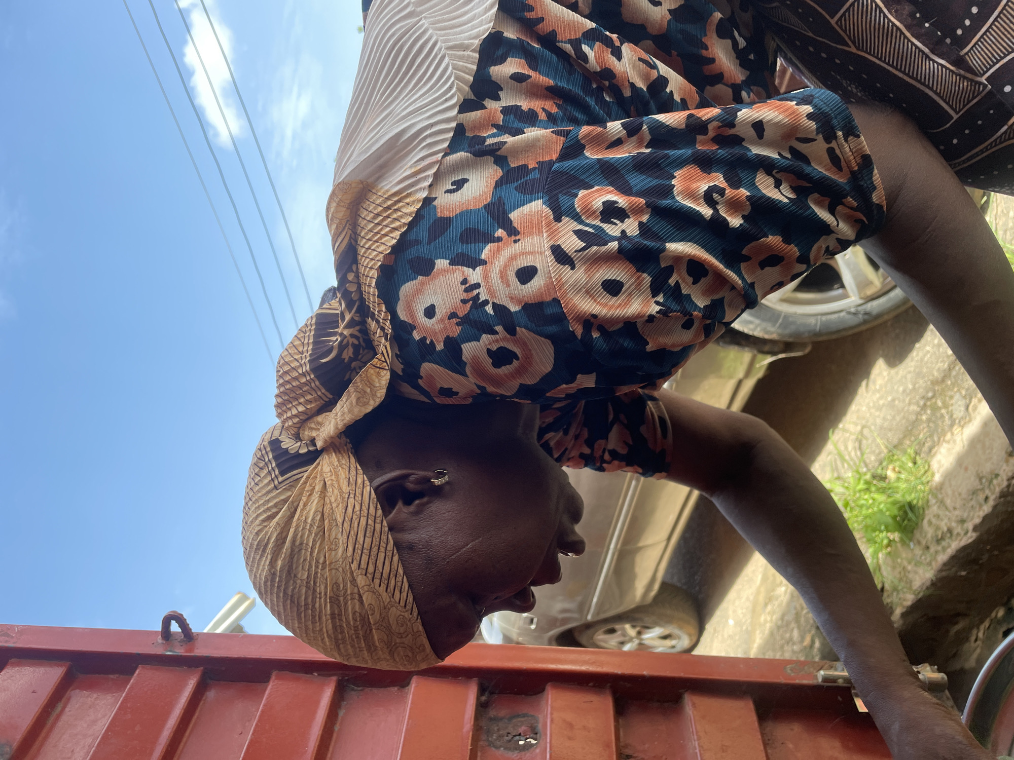 A dark-skinned woman wearing a patterned headscarf and floral-print top bends over a red, corrugated metal container. Her face is close to the container, and she appears to be examining or working on it. A vehicle, possibly a car, is visible in the background along with some vegetation. Blue sky and power lines can be seen in the upper left corner. The location is Jos, Nigeria.