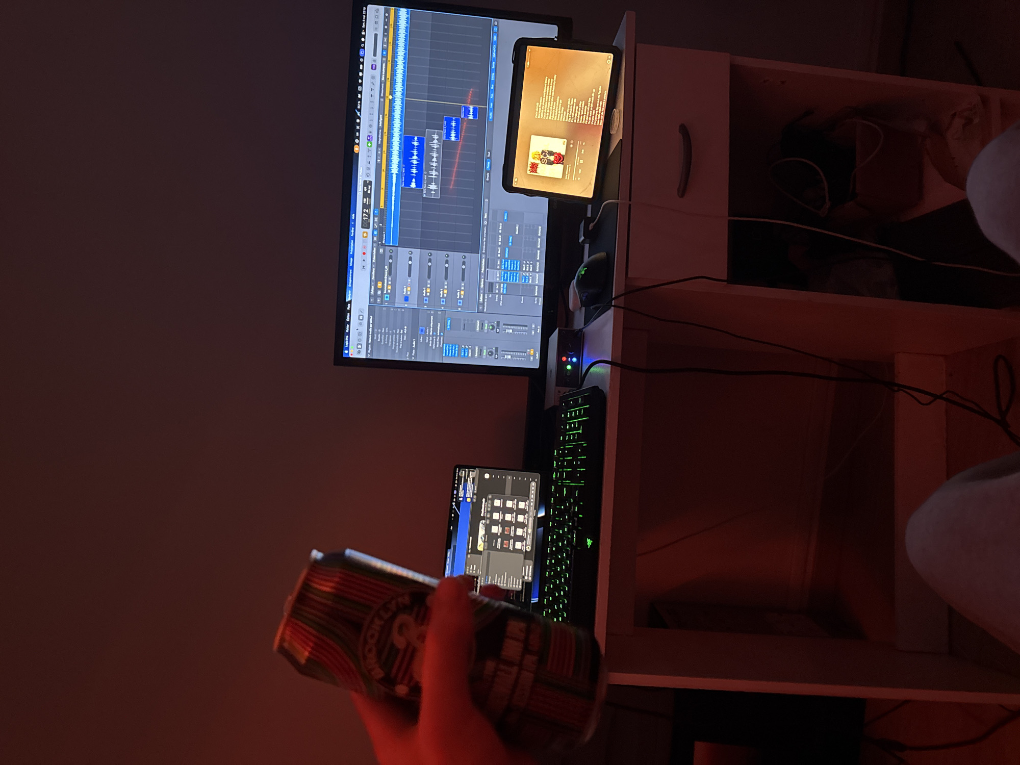 The image shows a desk setup in a room with a reddish lighting. On the desk are a large computer monitor displaying a digital audio workstation (DAW) interface, a tablet resting on the desk, a smaller monitor displaying a file explorer interface, a green glowing keyboard, and other computer accessories. A hand is holding a can of beer in the foreground. The desk has a shelf unit attached to it. The city of Paris, France is not depicted in this image.