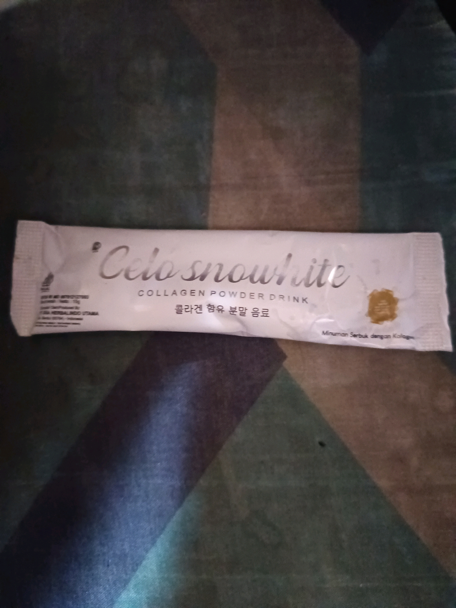 The image shows a single sachet of "Celo snowhite" collagen powder drink, likely found in a residential setting in South Jakarta, Indonesia. The sachet is white with a silver script logo and text. The background is a dark, textured surface, possibly a carpet or mat, with geometric patterns and lighting that creates shadows. There are no people or actions visible in the image, only the product itself placed on the surface.