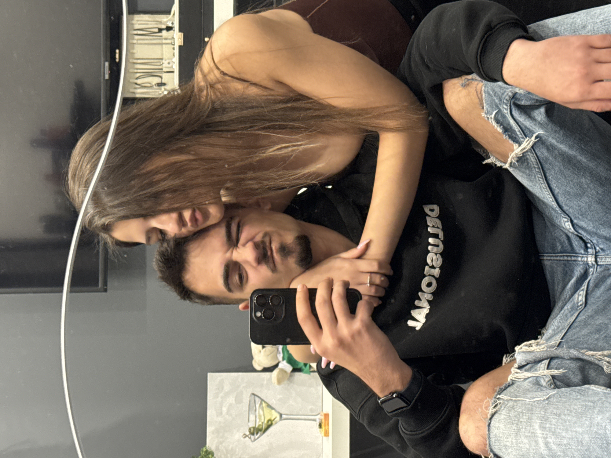 A couple takes a casual mirror selfie indoors, likely in a home in Sofia, Bulgaria. The man, with a goatee and mustache, holds a black smartphone (an iPhone 14 Pro or Pro Max) to capture the reflection. He wears a black hooded sweatshirt with the reversed text "BALENCIAGA" in white letters across the chest, and distressed light blue jeans with a tear exposing his knee. A black Apple Watch is visible on his left wrist. He makes a playful, winking expression.
The woman, with long brown hair, leans intimately against him, resting her head near his shoulder. She wears a dark top and has light pink manicured nails, with a ring visible on her finger. Her expression appears soft and affectionate.
The background, visible in the mirror's reflection, includes a dark-colored television screen on the left. Above it, a shelf or cabinet contains various small items, possibly jewelry or electronic components. Below the couple, a framed artwork featuring a martini glass with green olives is displayed, alongside a partial view of a small white teddy bear. The lighting suggests an indoor setting with artificial light. The overall mood is casual and intimate.