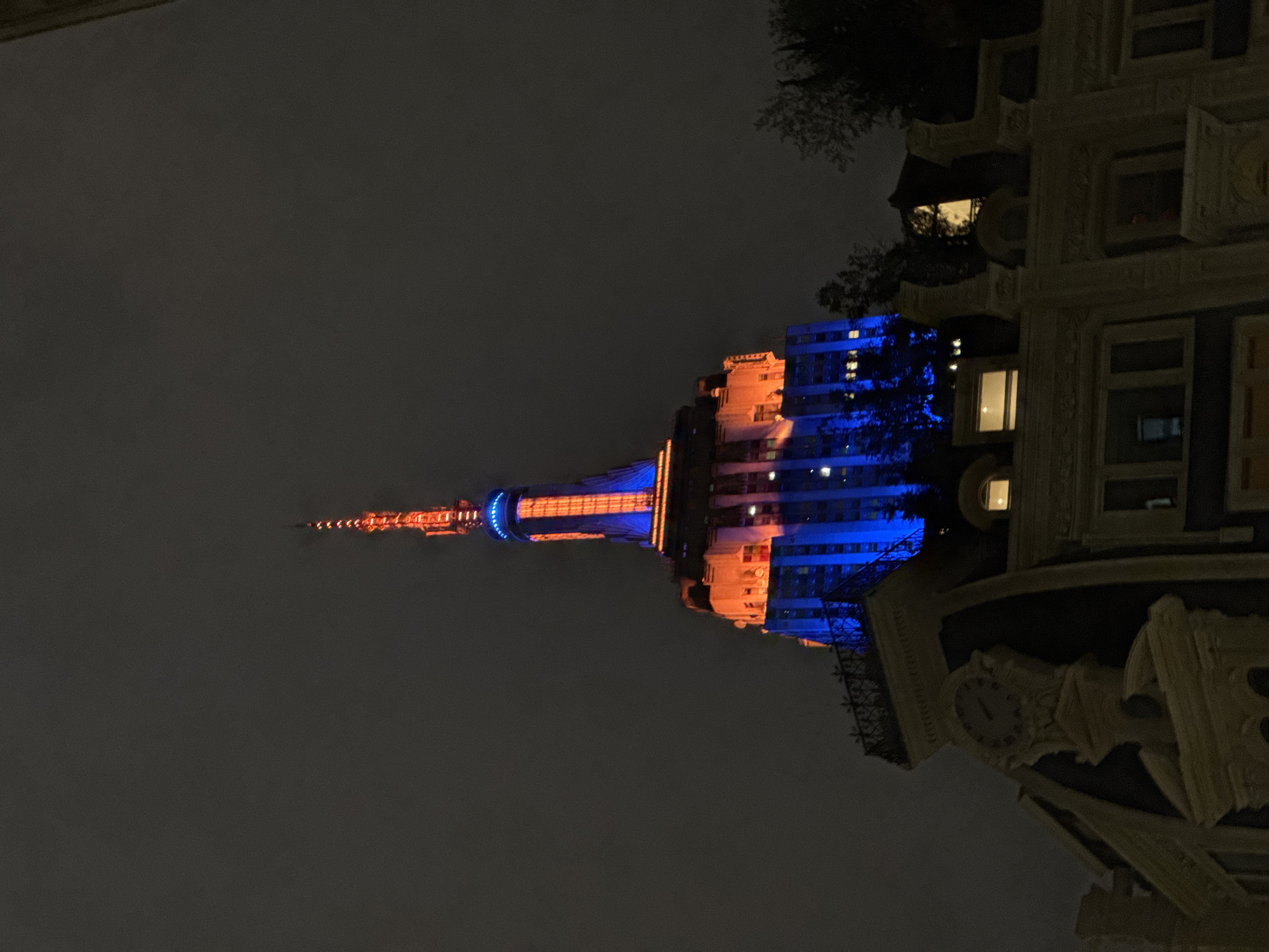 The image shows the New York Life Building, located in New York City, United States of America, illuminated in orange and blue lights against a dark, cloudy sky at night. Part of another building, possibly the Grand Central Terminal, is visible on the right side of the image. The clock on this building appears to be illuminated. The scene seems to be taken from street level.