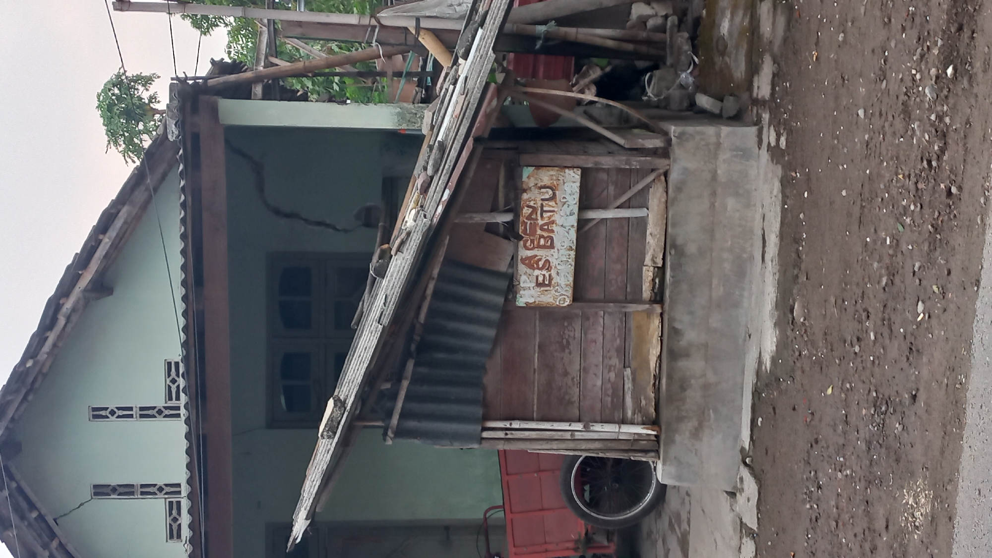 This image captures a weathered, wooden kiosk with a corrugated tin roof, situated in front of a pale green house with visible cracks. A red cart with a large wheel is parked to the left of the kiosk. The kiosk itself displays a sign with the words "AGEN ES BATU" which translates to "Ice Agent". The ground in front of the kiosk is unpaved and appears to be a mix of dirt and gravel. The overall scene suggests a humble, possibly rural, setting in the city of Pare, Indonesia, a region known for its agricultural prominence and a local culture that often involves small, roadside businesses. The condition of the house and the kiosk, along with the unpaved ground, point to a location that is not in a highly developed urban area.