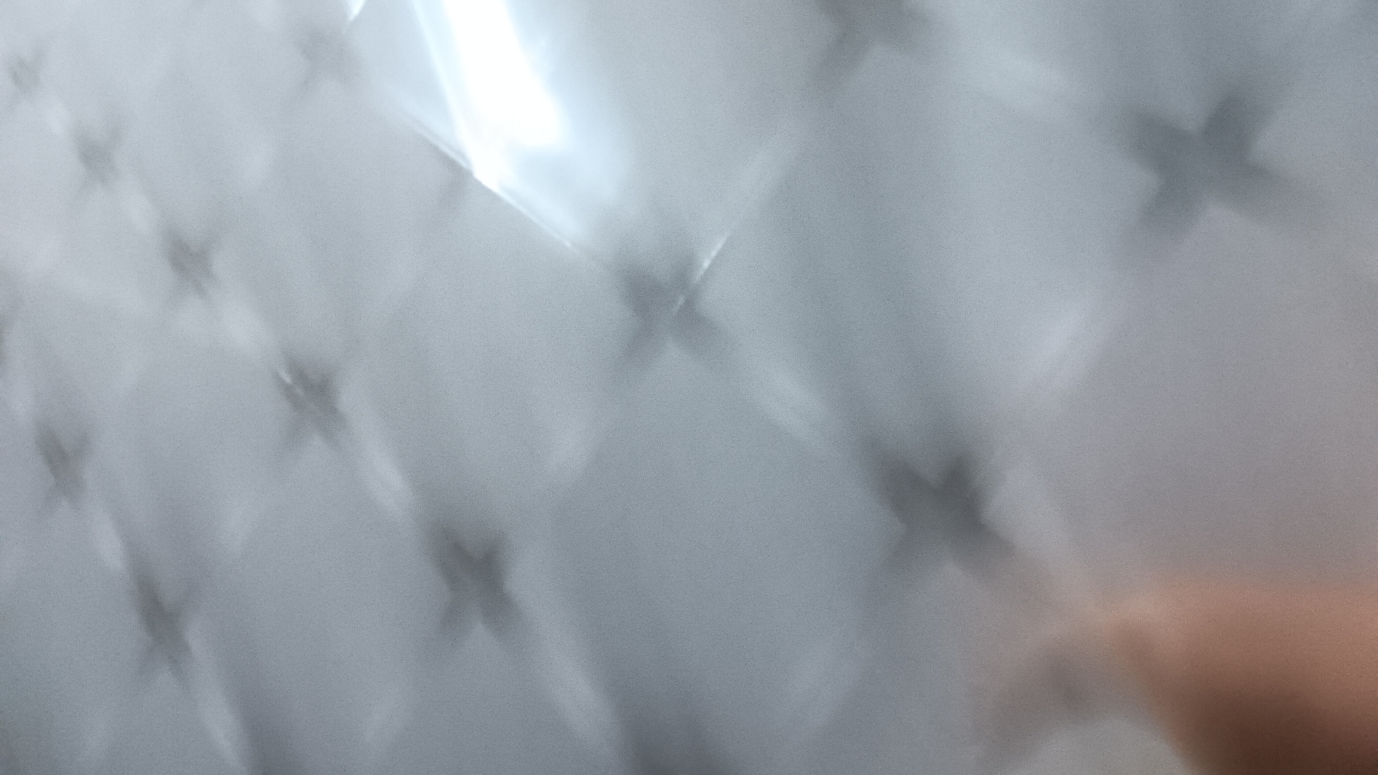 The image shows a blurry, abstract pattern of intersecting lines and shapes that resemble a grid of crosses. The lighting is diffuse and creates a soft, ethereal effect, with a bright light source in the upper right quadrant of the image. There is a hint of a human hand or arm in the lower left corner, appearing as a soft, flesh-toned blur against the grey background. The overall impression is one of diffused light and shadow, with a subtle sense of depth. The image is too abstract and lacks sufficient detail to identify any specific location, including Sonobekel, Indonesia.