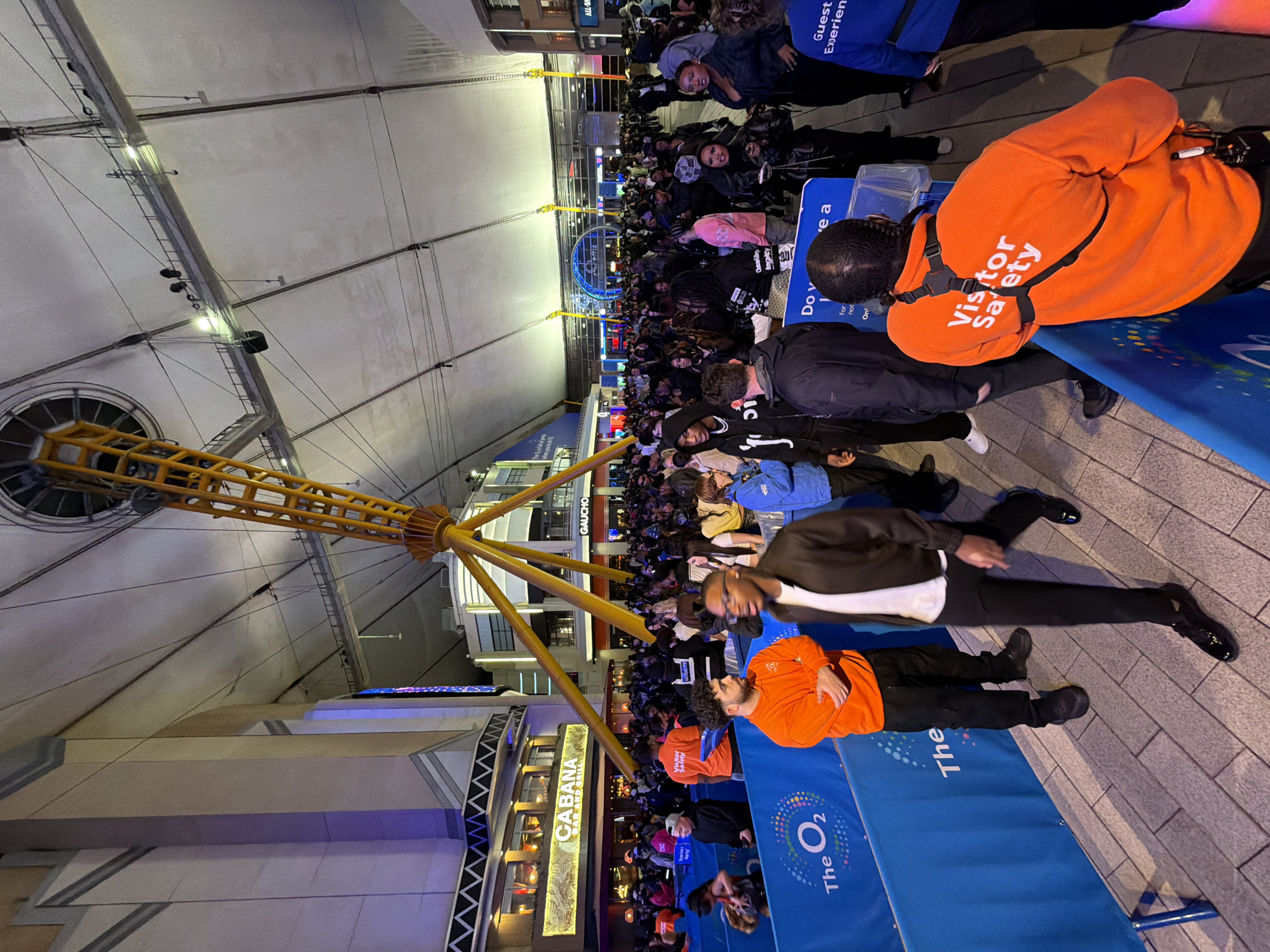 The image depicts a bustling scene at the O2 Arena in London, United Kingdom. The iconic dome structure of the O2 is visible, with its distinctive yellow support tower. A large crowd of people is gathered, illuminated by the arena's lighting and surrounding buildings. Several individuals wearing orange "Visitor Safety" vests are present, likely event staff. The "Cabana Bar and Grill" is visible, as is signage for "The O2". The crowd appears to be waiting or attending an event.