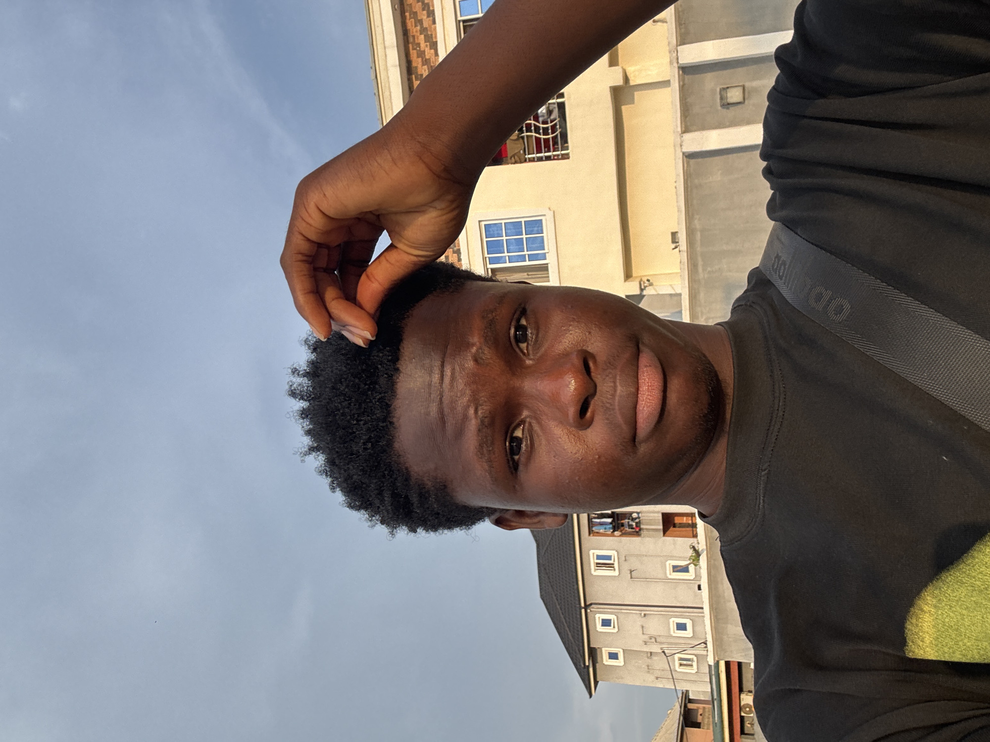 A young Black male is pictured in a close-up selfie outdoors, with a building and cloudy sky in the background. The male is wearing a black t-shirt and has his right hand raised, touching the top of his head. He has dark, curly hair and is looking directly at the camera with a neutral expression. The building in the background appears to be a multi-story residential structure. The sky is a mix of blue and gray, suggesting an overcast or partly cloudy day. The overall lighting suggests it is daytime. The location is identified as Ihiagwa, Nigeria.