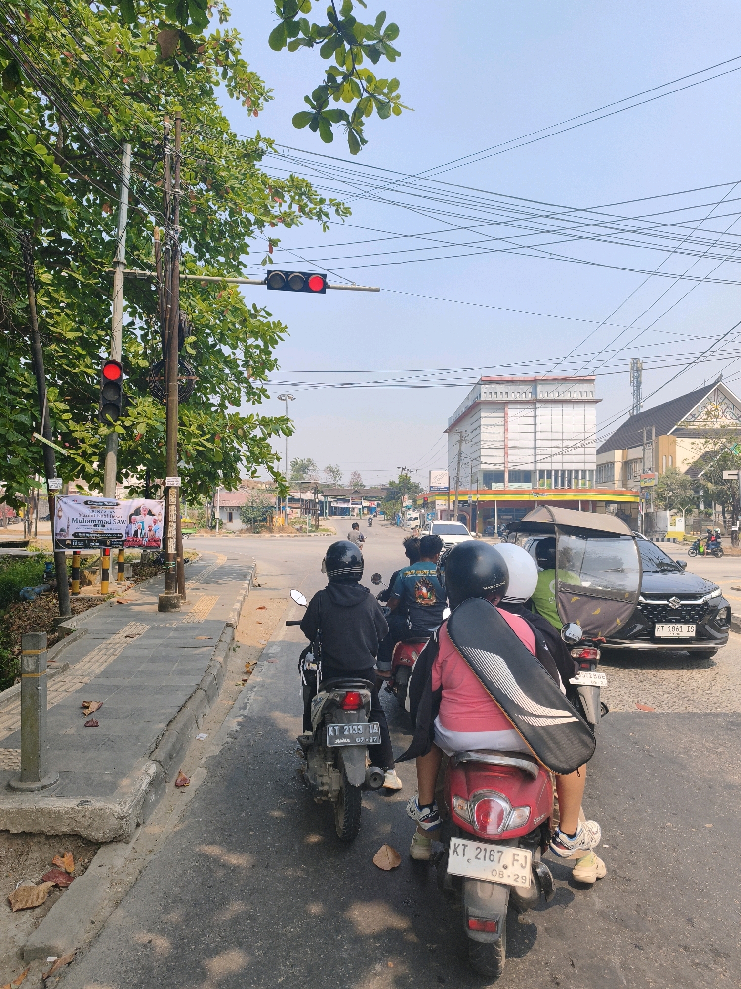 The image captures a street scene in Samarinda, Indonesia, where several people are on motorcycles at a traffic light. Two traffic lights are visible, both displaying red, indicating that vehicles should stop. In the foreground, three motorcycles are lined up, with riders wearing helmets. One motorcycle is red, another is black, and the third is partially obscured. The rider of the black motorcycle in the middle is wearing a black hoodie, and the rider of the red motorcycle in the foreground is wearing a pink shirt and a black strap across their back. A black Suzuki car is stopped behind the motorcycles, and a three-wheeled vehicle with a canopy is also present. The street is paved, with a sidewalk to the left featuring a pedestrian crossing marked by yellow tactile paving. A traffic sign and a banner with Indonesian text are mounted on a pole near the sidewalk. Trees with green leaves line the left side of the street, and utility wires crisscross overhead. Buildings are visible in the background, including a modern, multi-story building with white and red accents. The sky is clear and blue.