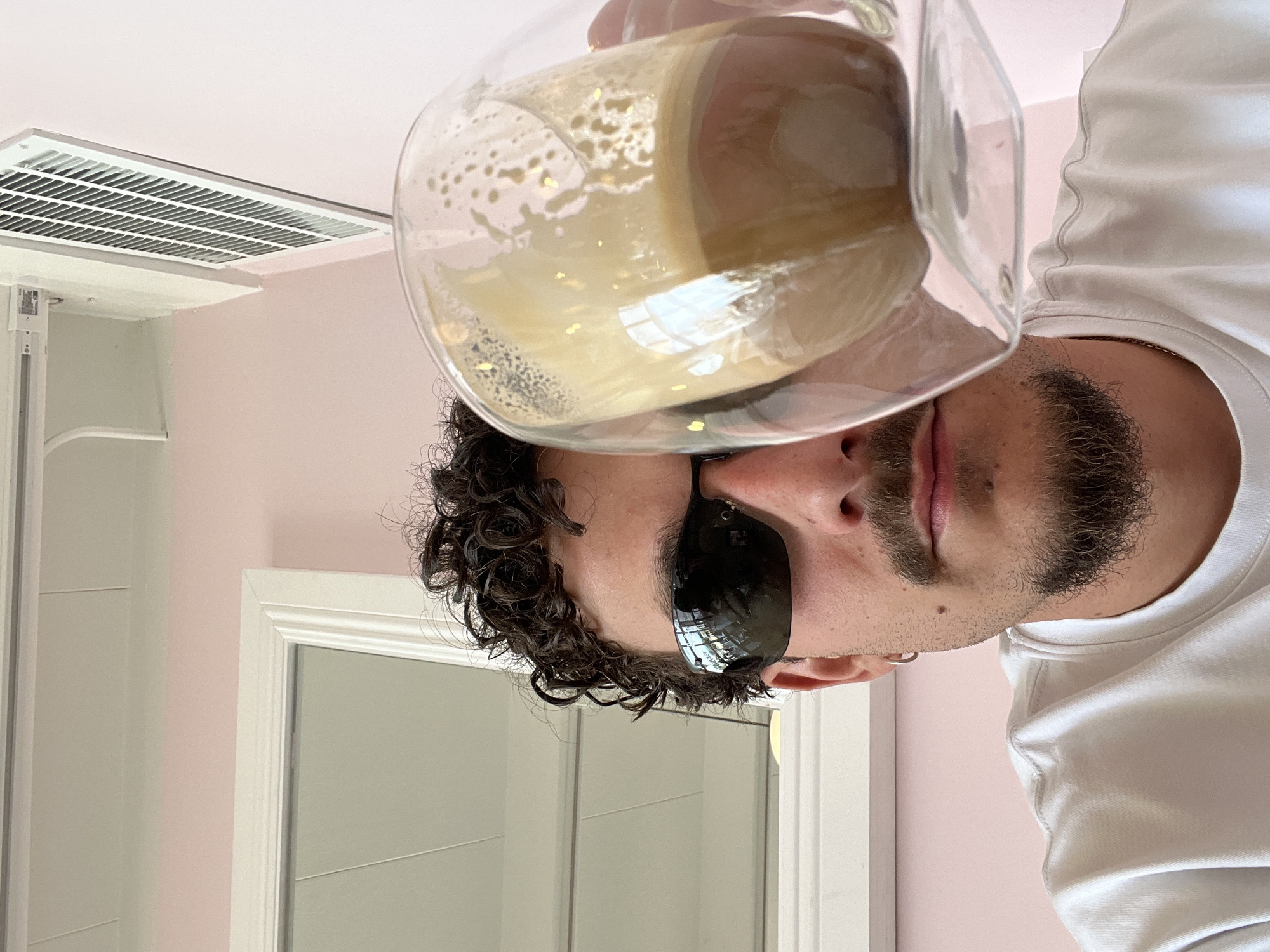 A man in sunglasses and a white t-shirt holds a glass of liquid in front of his face in an indoor setting with pink walls. He has curly hair and a beard. In the background, there is a vent and a mirror or door with a white frame. The location is likely a bathroom, as indicated by the mirror and vent. The exact location in Palm Desert, United States of America is unknown.