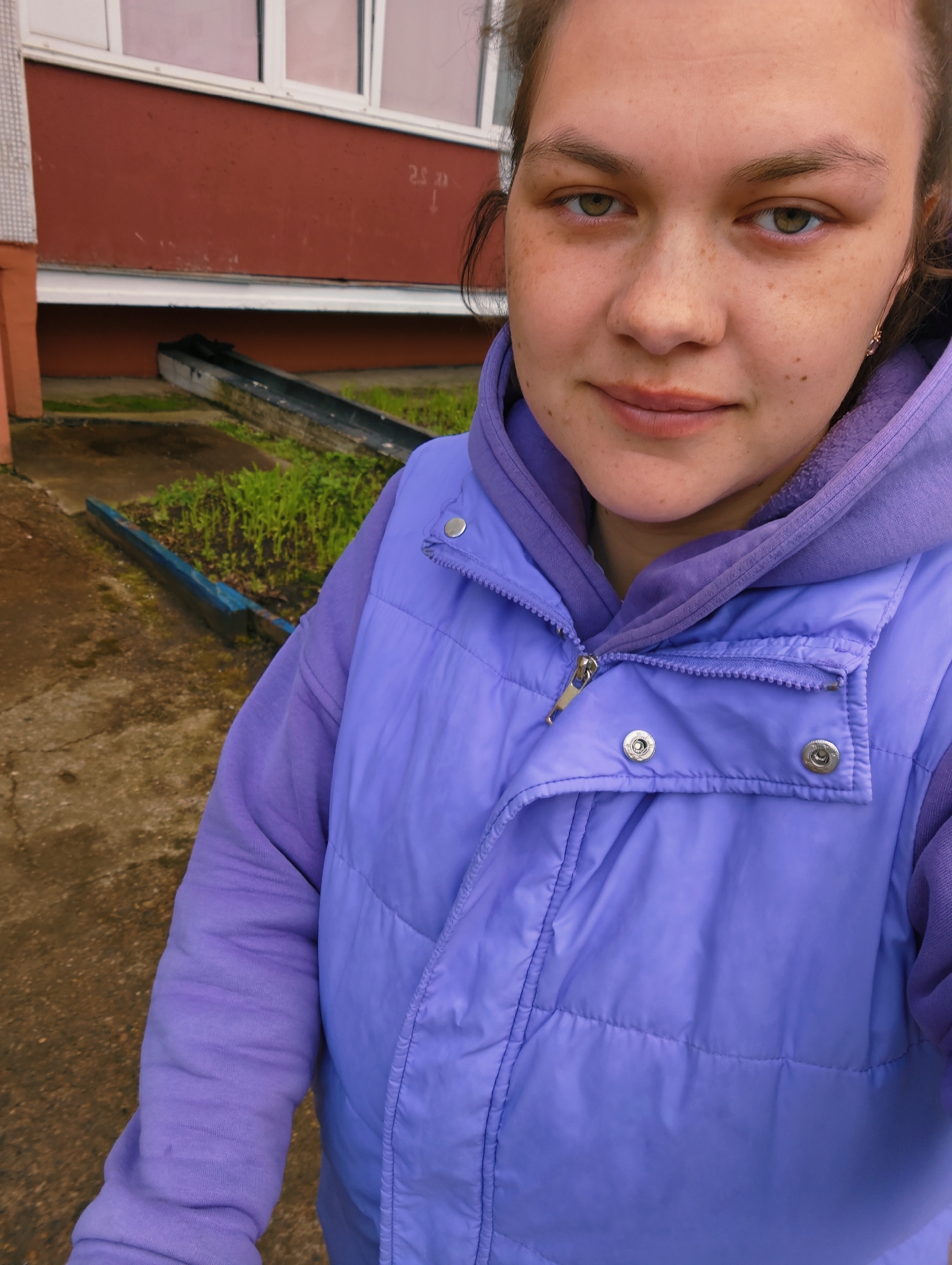 A young woman is captured in a medium close-up selfie outdoors, likely in Chaikovsky, Russia. She has light skin, visible freckles, and light-colored eyes (green or hazel). Her dark hair is pulled back, and she wears a purple hooded sweatshirt layered under a lighter purple puffer vest, which is partially unzipped. A small, gold earring with a pink stone dangles from her right ear. She looks directly at the camera with a calm, neutral expression and a faint smile.
The background reveals a damp concrete ground, and a red-brown building wall with a white-framed window. Below the window, faint white text, possibly "2.5 >" or "25 >", is visible on the wall, accompanied by a downward-pointing arrow. On the ground, a long, dark wooden trough-like structure, possibly a planter or drainage channel, contains small green sprouts. Another blue painted wooden plank lies near it. The scene suggests an outdoor, possibly residential or utilitarian area, under what appears to be an overcast sky, indicated by the soft, diffused lighting.