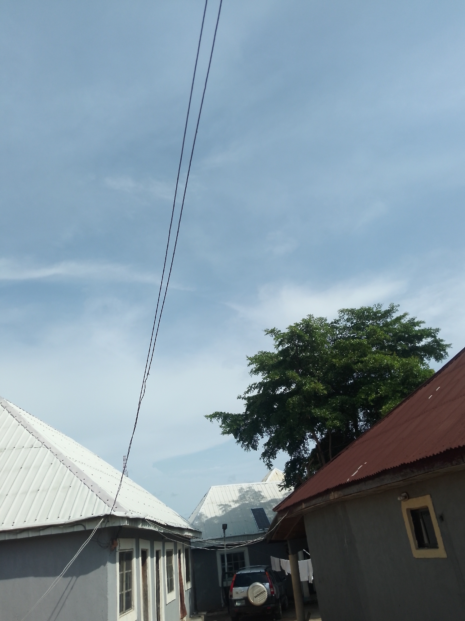 The image, taken in Serti, Nigeria, depicts a residential area under a pale blue sky with scattered white clouds. In the foreground, two houses are visible. The house on the left has a light gray exterior and a white corrugated metal roof. It features several windows with wooden frames and is partially obscured by a power line. The house on the right has a dark gray exterior and a reddish-brown corrugated metal roof. It has a small, dark-colored window with a yellow frame. A large, leafy green tree stands between the two houses. In the background, another house with a white roof and a dark solar panel is visible. A silver SUV is parked in front of the house on the right. White laundry hangs on a clothesline between the houses on the right. The scene suggests a typical day in a residential neighborhood.