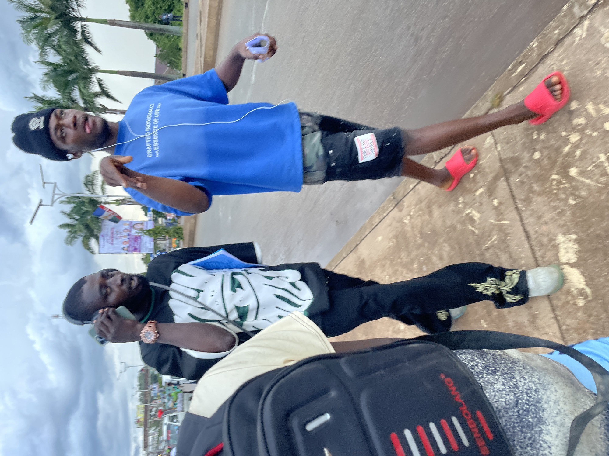 Two young men walk side-by-side on a paved sidewalk in Jos, Nigeria. The man on the left, wearing a black and white t-shirt, a watch, and headphones, holds a mobile phone to his ear. The man on the right, in a blue t-shirt and camouflage shorts, wears a black beanie and earbuds, and appears to be speaking while gesturing with his right hand. A third person, carrying a black backpack, is partially visible in the foreground on the left. The background shows a street with vehicles and palm trees, under a cloudy sky.