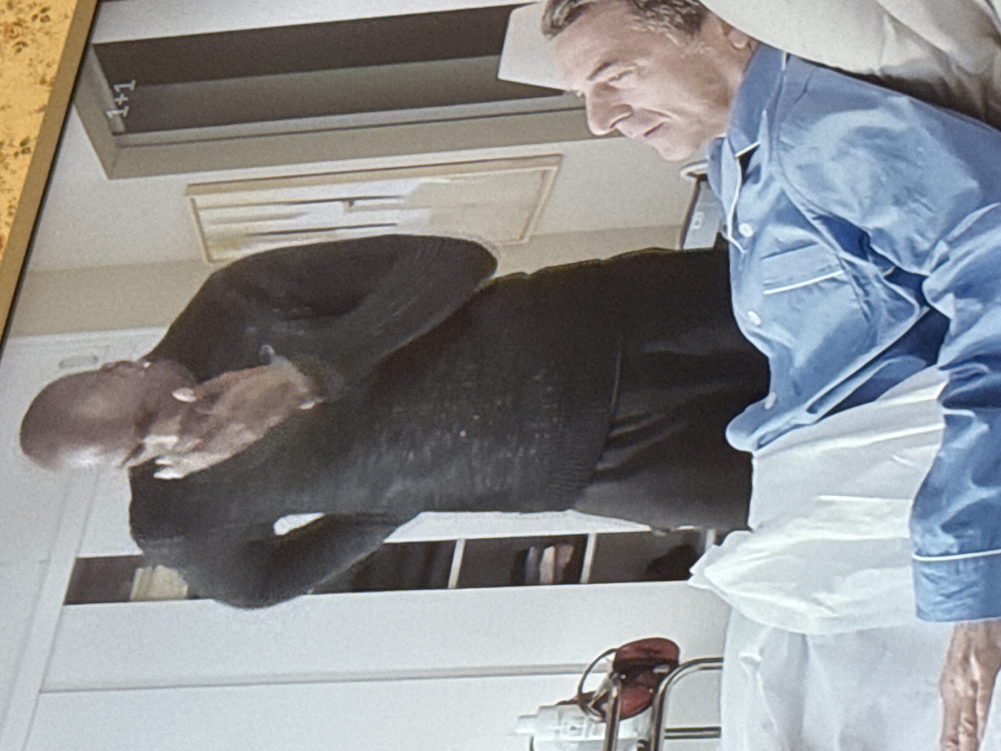 Two men are visible in the scene. One man, seen in profile from the shoulders up, is wearing blue pajamas. The other man is wearing a black sweater. The man in the black sweater is bald and holding his hands near his face. The scene appears to be taking place in a hospital room. There are medical devices and white linens visible. The location is not possible to determine from this image.