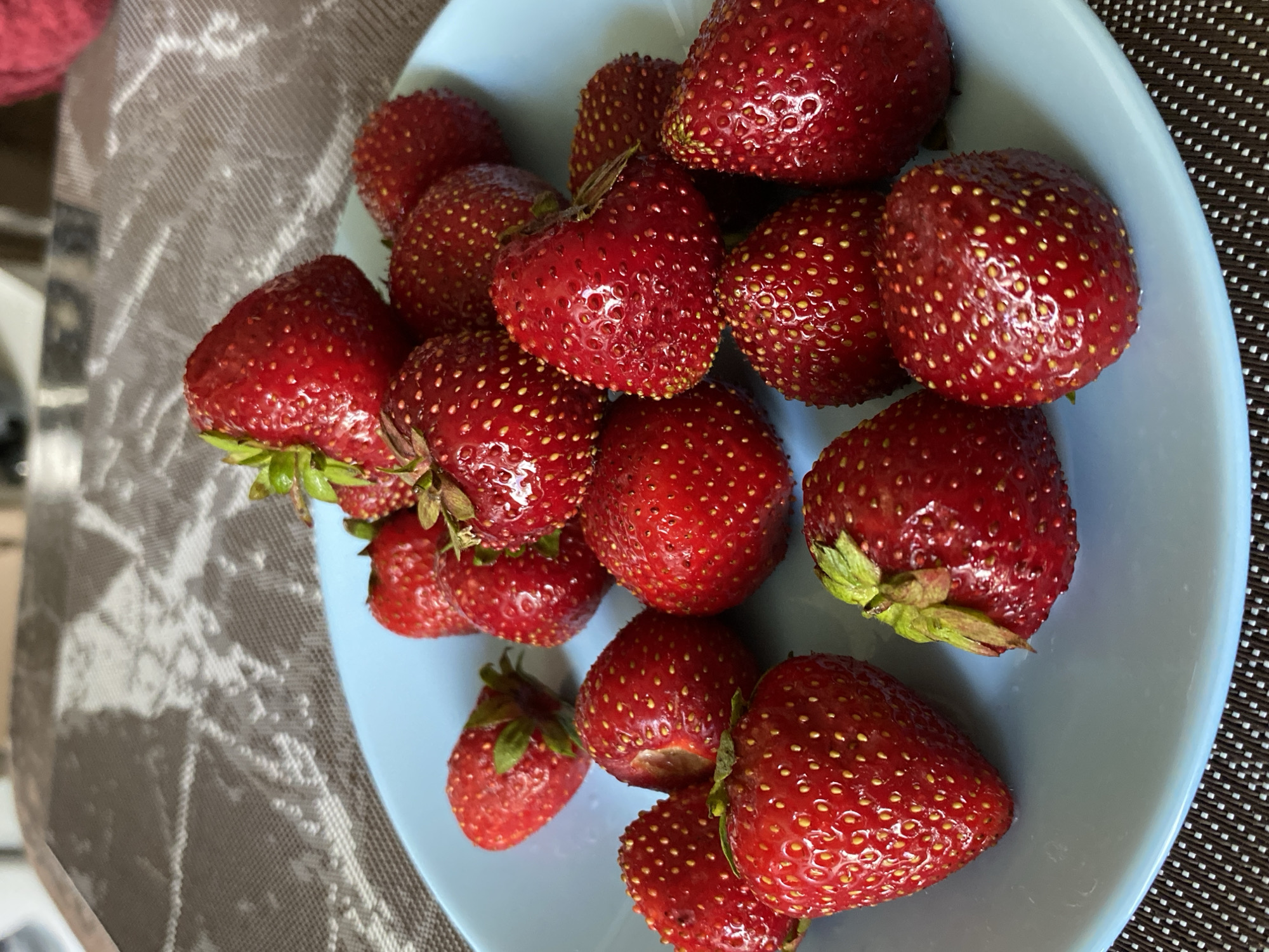 A close-up, overhead shot shows a light blue plate filled with ripe strawberries. The strawberries are bright red with visible seeds and green stems. The plate rests on a patterned placemat with a dark brown base and a white diamond pattern. In the background, a blurred surface with a textured pattern, possibly a tablecloth, is visible. The overall scene suggests a casual setting.