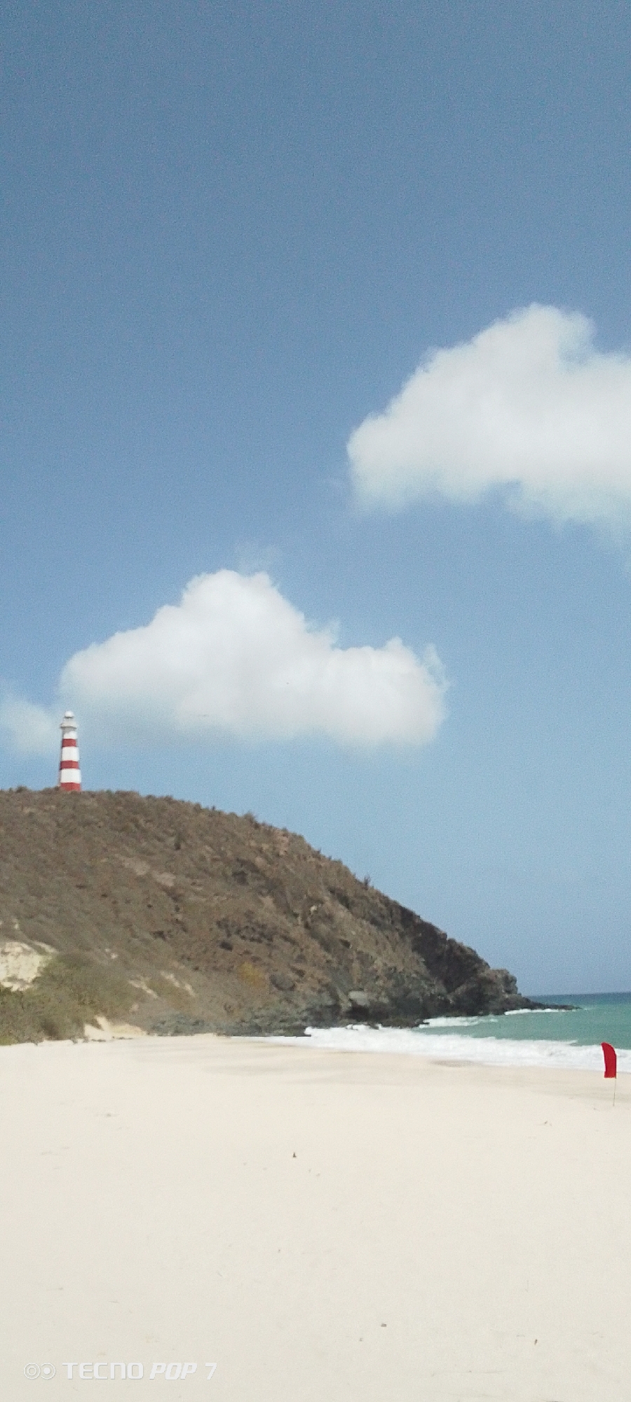 The image depicts a coastal scene in Pedro González, Venezuela. A red and white striped lighthouse stands atop a hill overlooking a sandy beach. The hill is covered in dry vegetation. The beach has a red flag planted near the shoreline, where gentle waves are visible. The sky is blue with scattered white clouds.